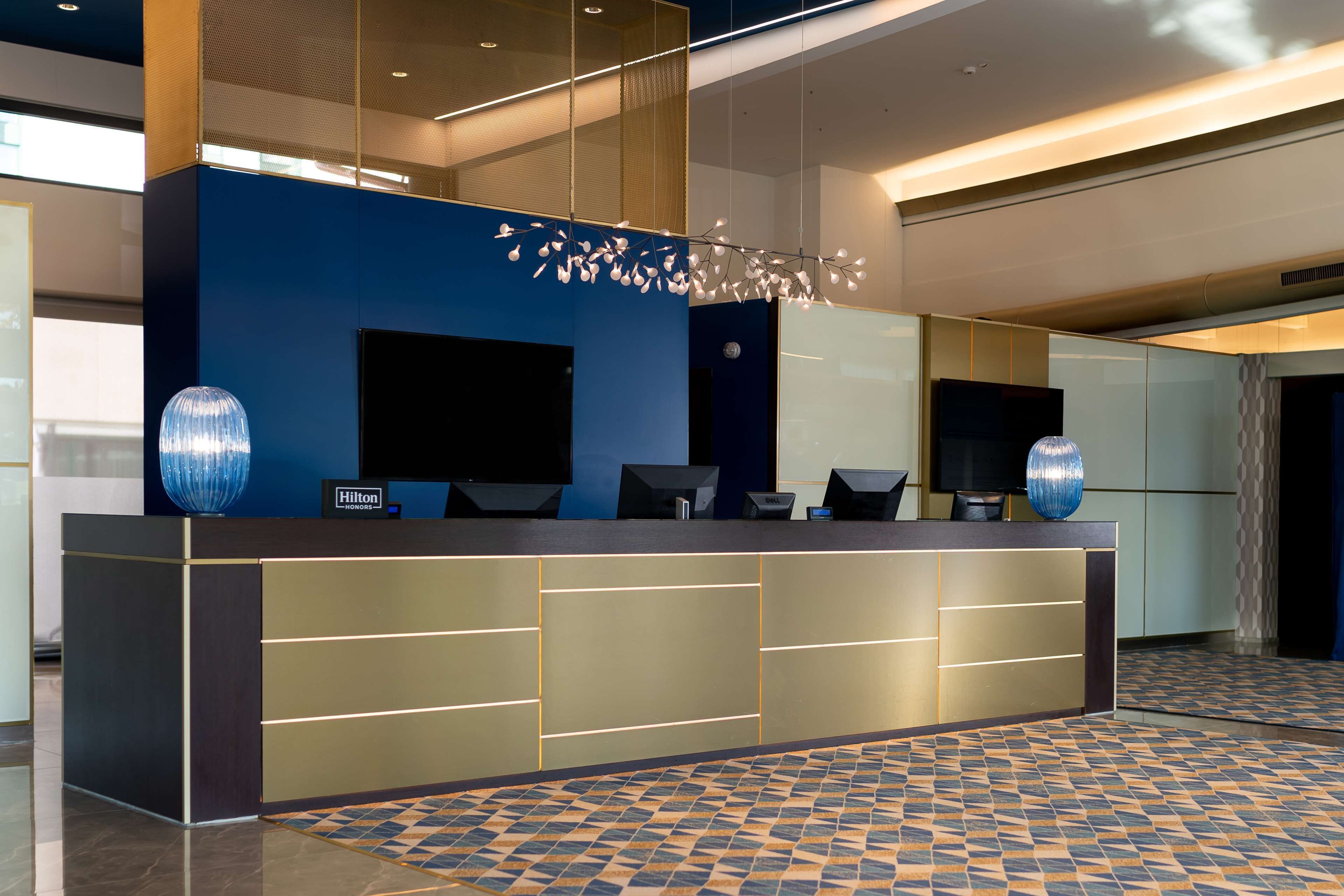 Zona de recepción de un hotel moderno con un muro azul decorativo, una pantalla digital y una lámpara ornamental.
