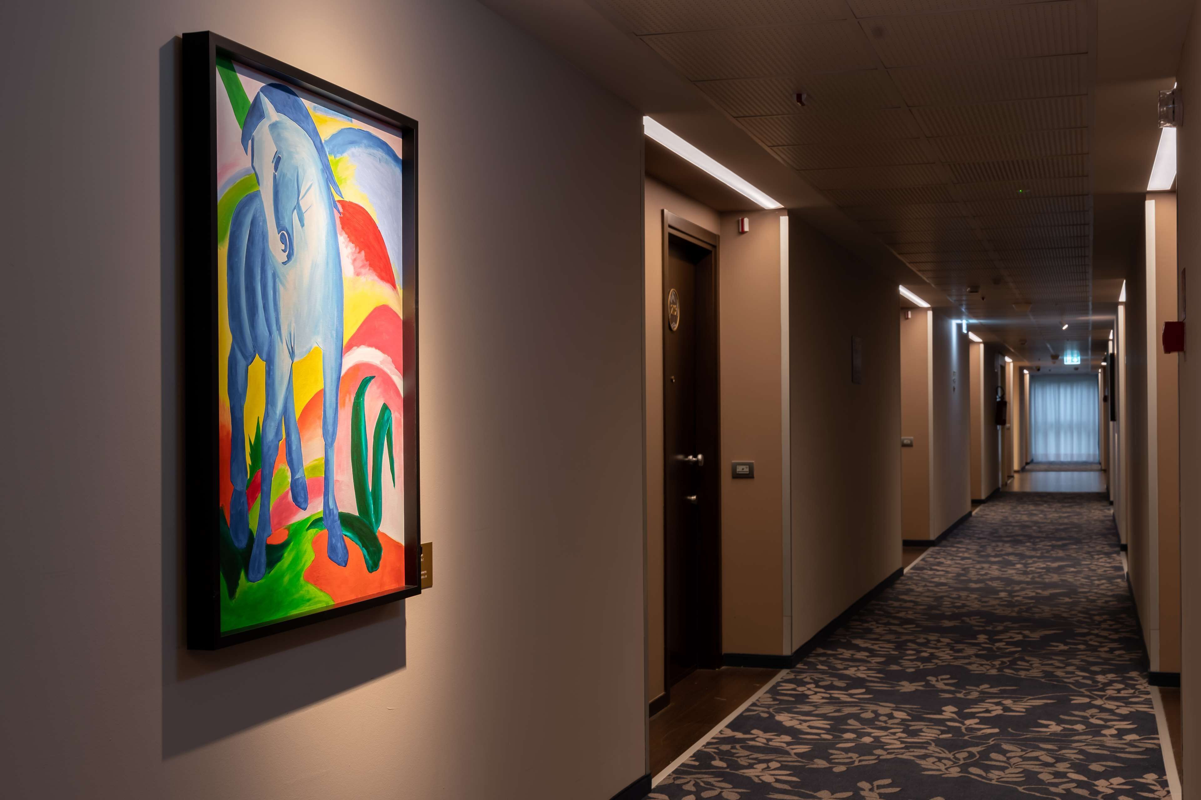 A hallway with a colorful abstract painting on the wall, a carpeted floor, and doors leading to rooms.