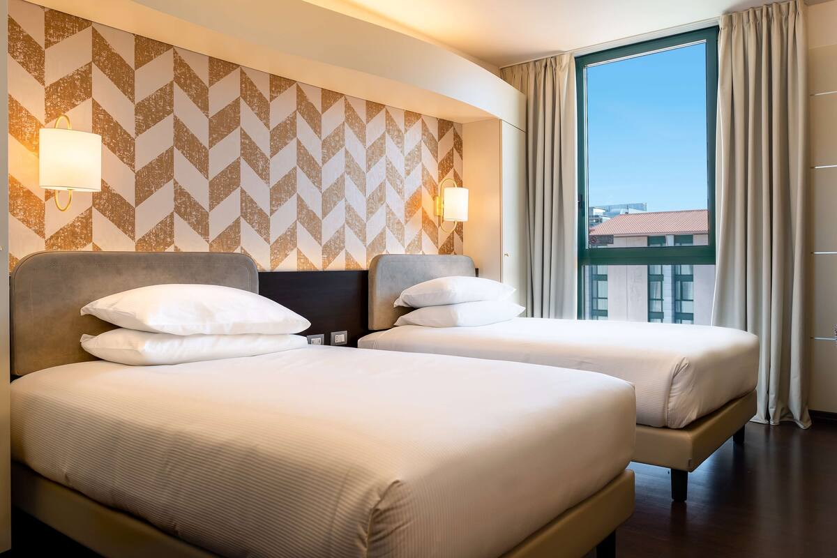 Habitación de hotel con dos camas, un ventanal con cortinas y un papel tapiz estampado.