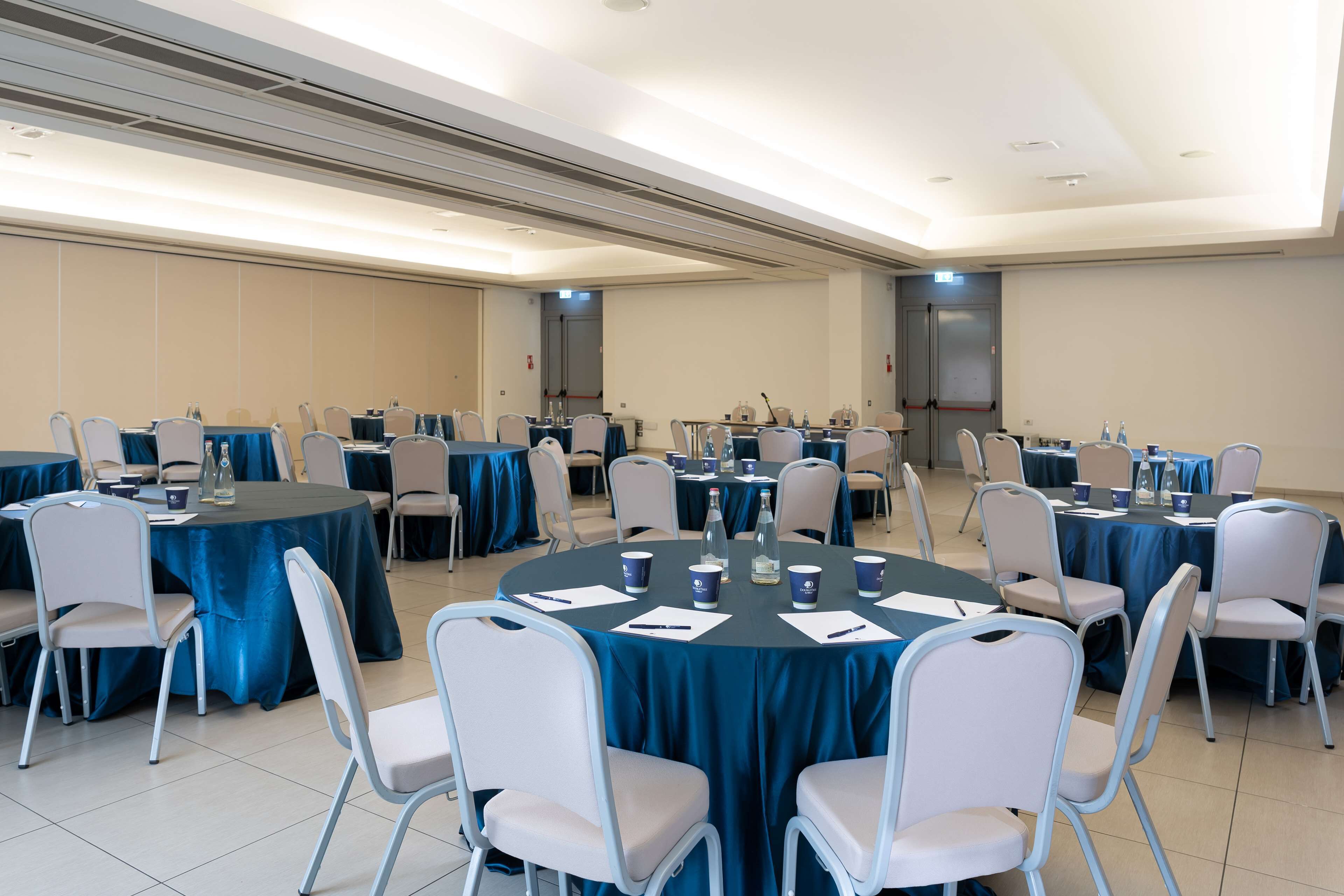 Un salón de conferencias con varias mesas redondas, cada una rodeada de sillas blancas. Las mesas están cubiertas con manteles azules, y hay botellas de agua y vasos sobre ellas.