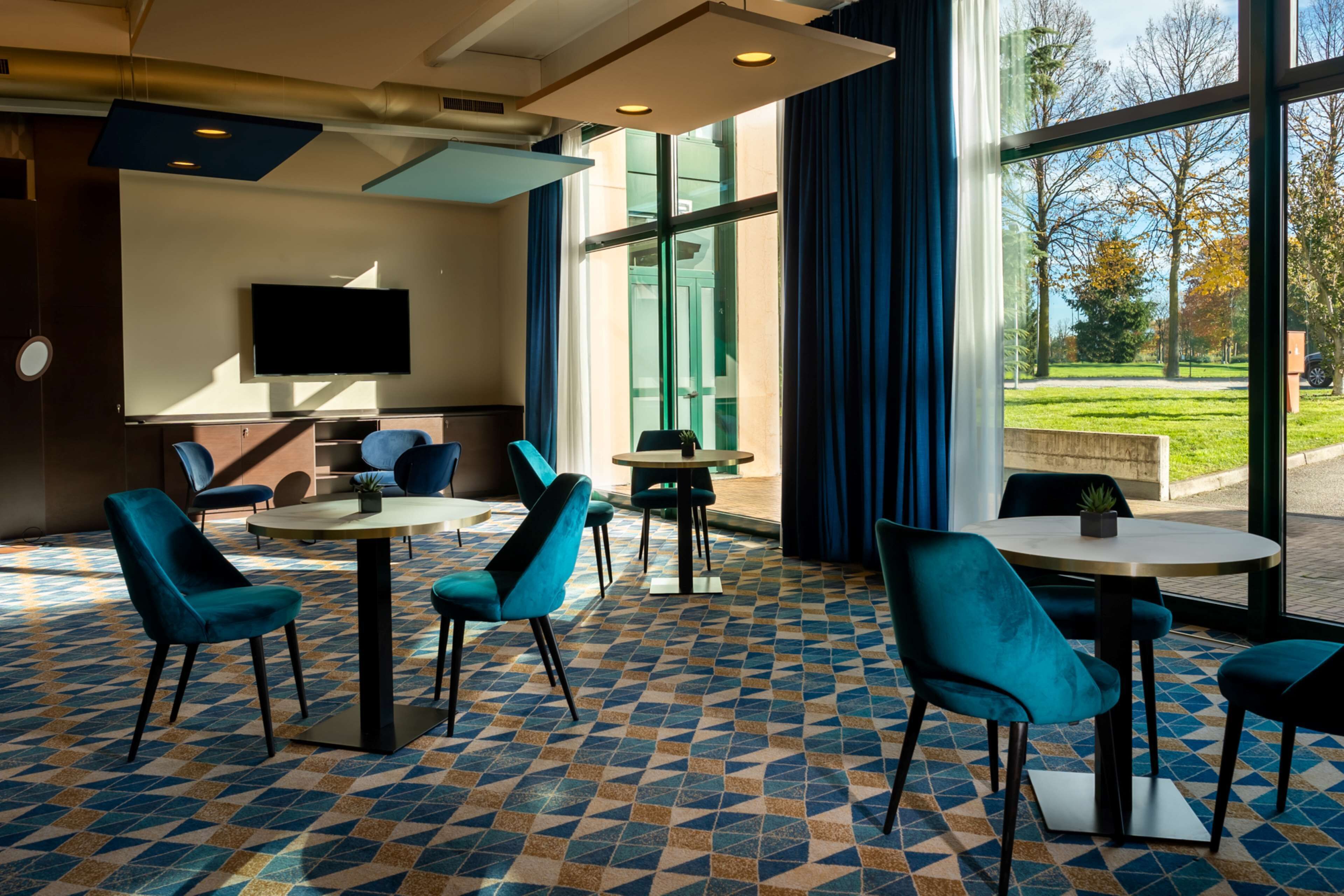 A modern hotel lobby with a geometric patterned floor, teal upholstered chairs, and a flat-screen TV mounted on the wall.