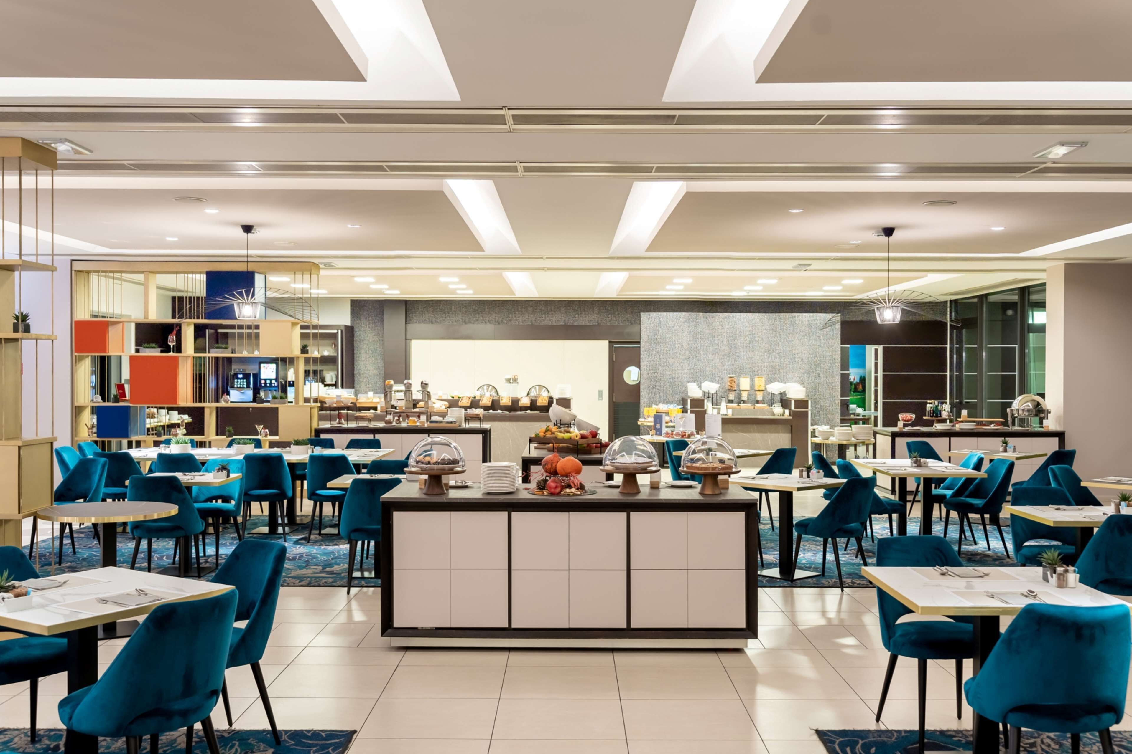 A modern hotel dining area with a central buffet station, multiple tables, and blue chairs.