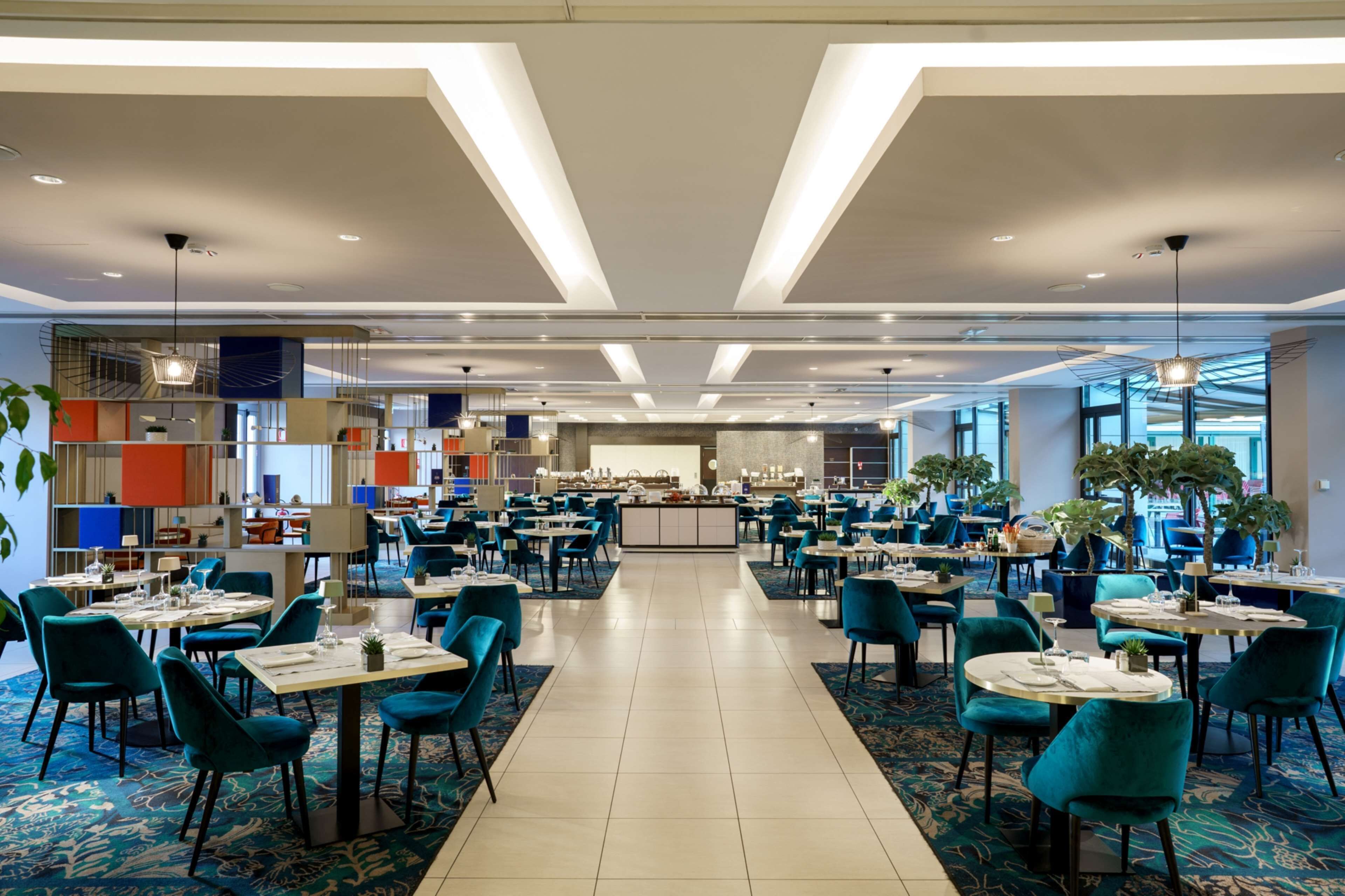 A spacious hotel dining area with multiple tables and chairs, a central counter, and large windows providing natural light.