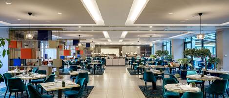Un comedor de hotel espacioso con varias mesas y sillas, una barra central y grandes ventanales que dejan entrar luz natural.