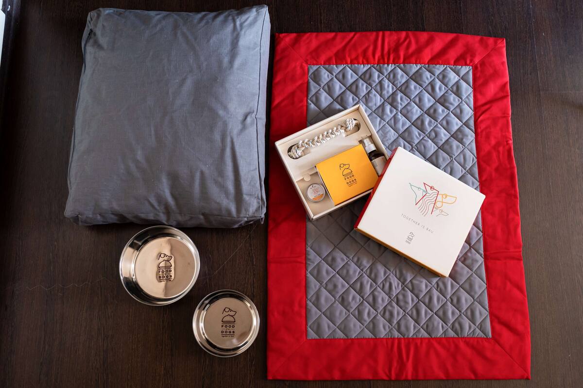 Una mesa ordenada con camino de mesa acolchado rojo y gris, un cojín gris, un recipiente de metal y una caja con logotipo y texto.