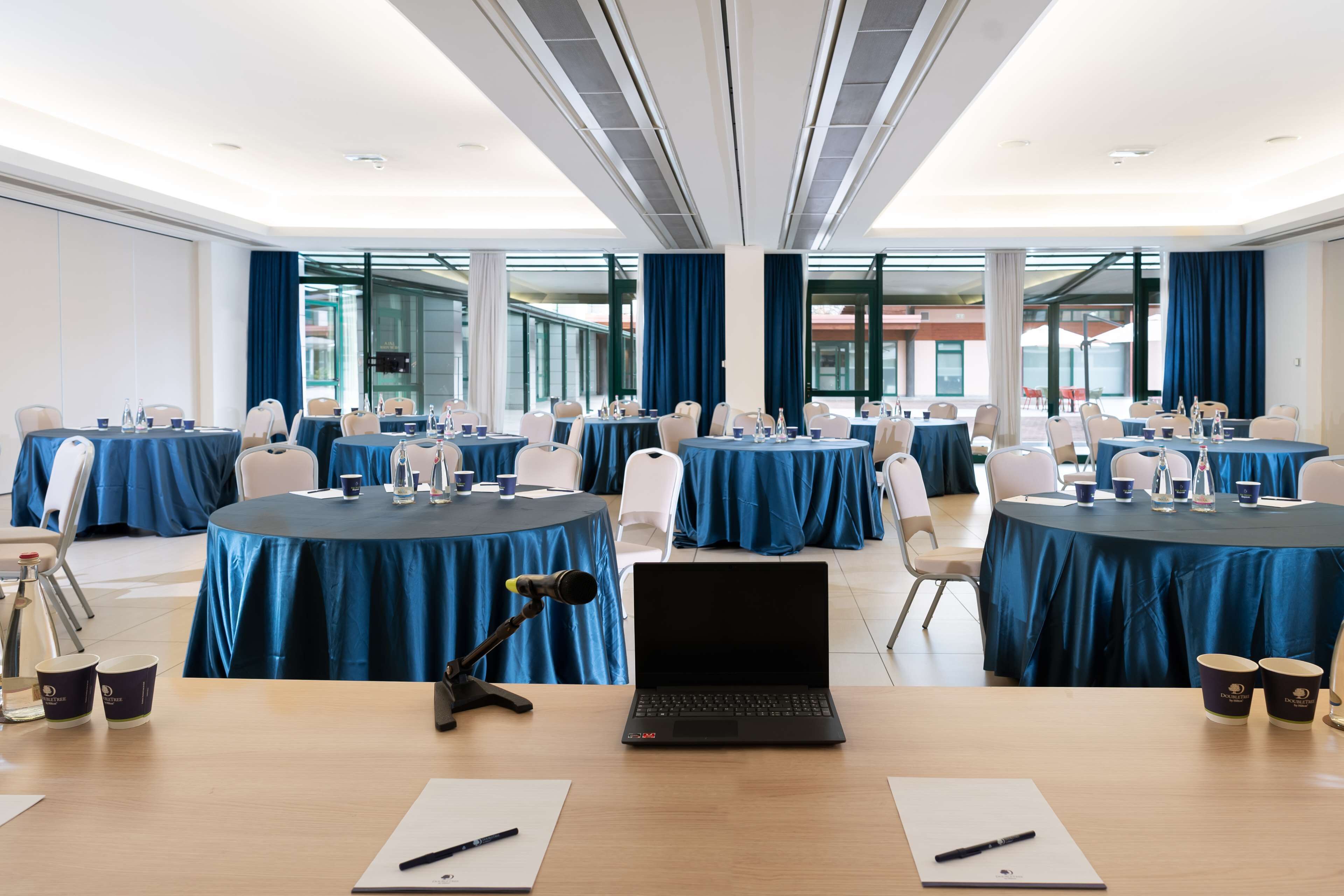 Un salón de conferencias con varias mesas, cada una cubierta con un mantel azul, equipado con computadoras portátiles, micrófonos y botellas de agua.