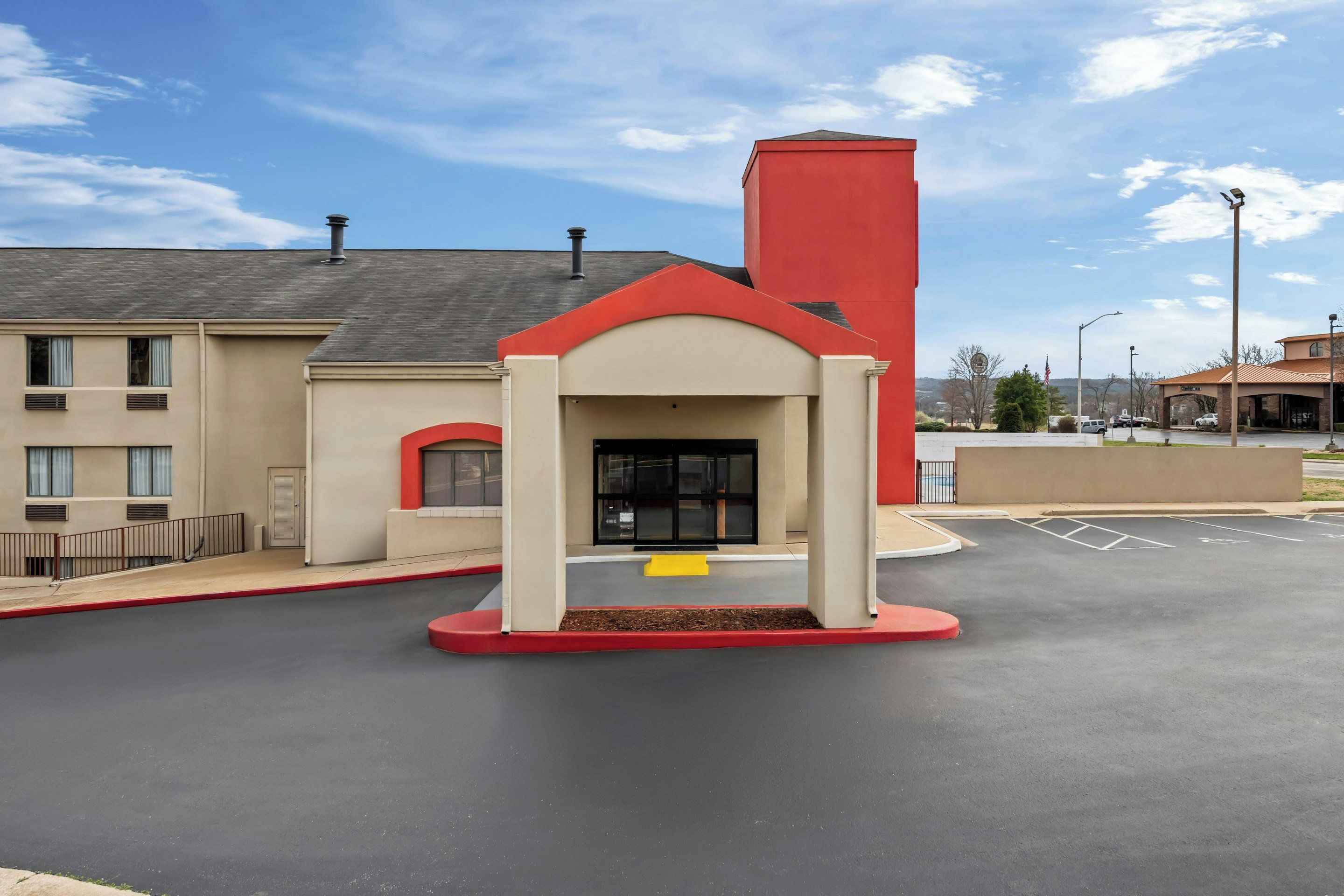 A hotel entrance with a red and beige facade, a parking lot, and a clear sky.