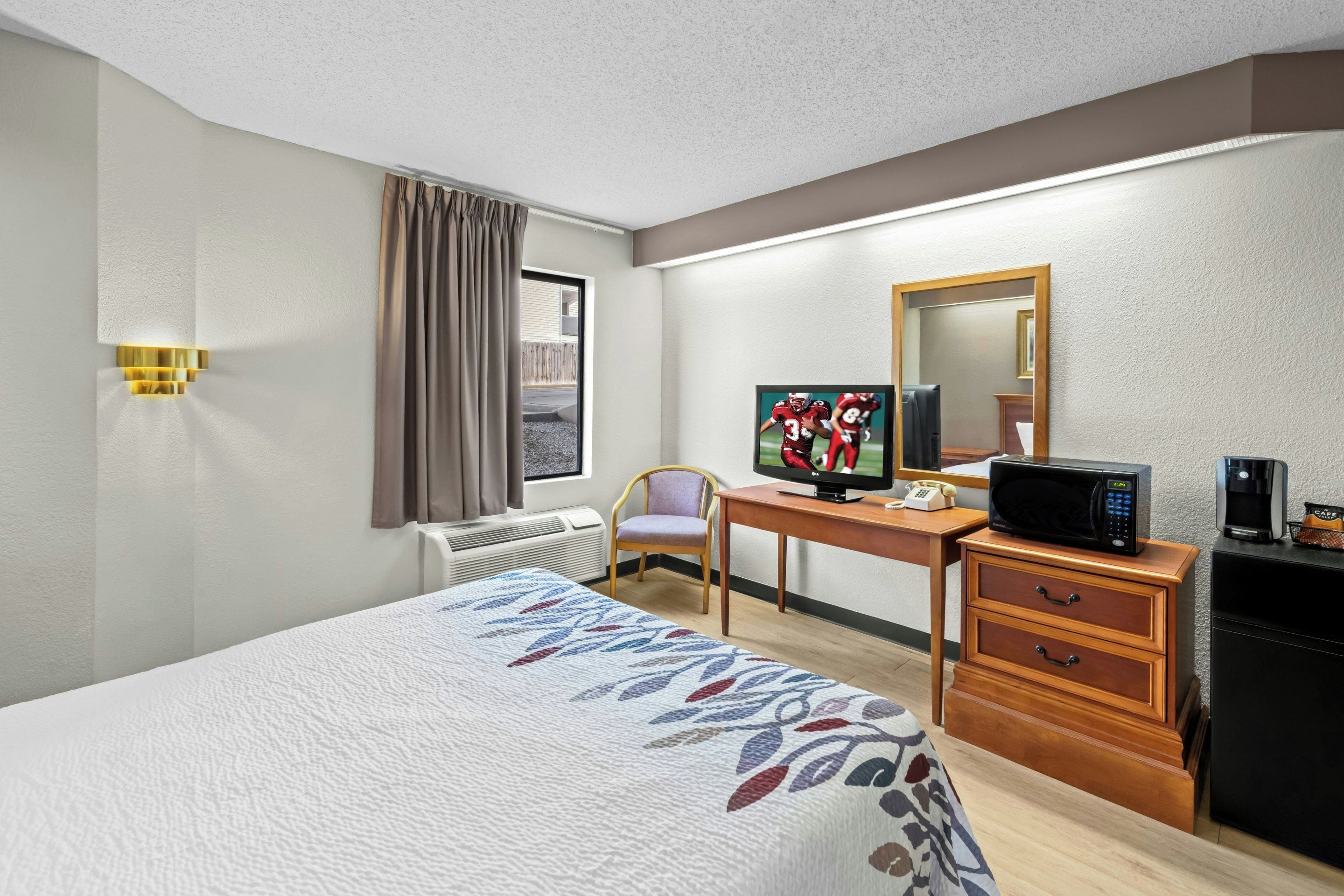 A hotel room with a bed, a desk with a computer, a microwave, a coffee maker, and a chair.