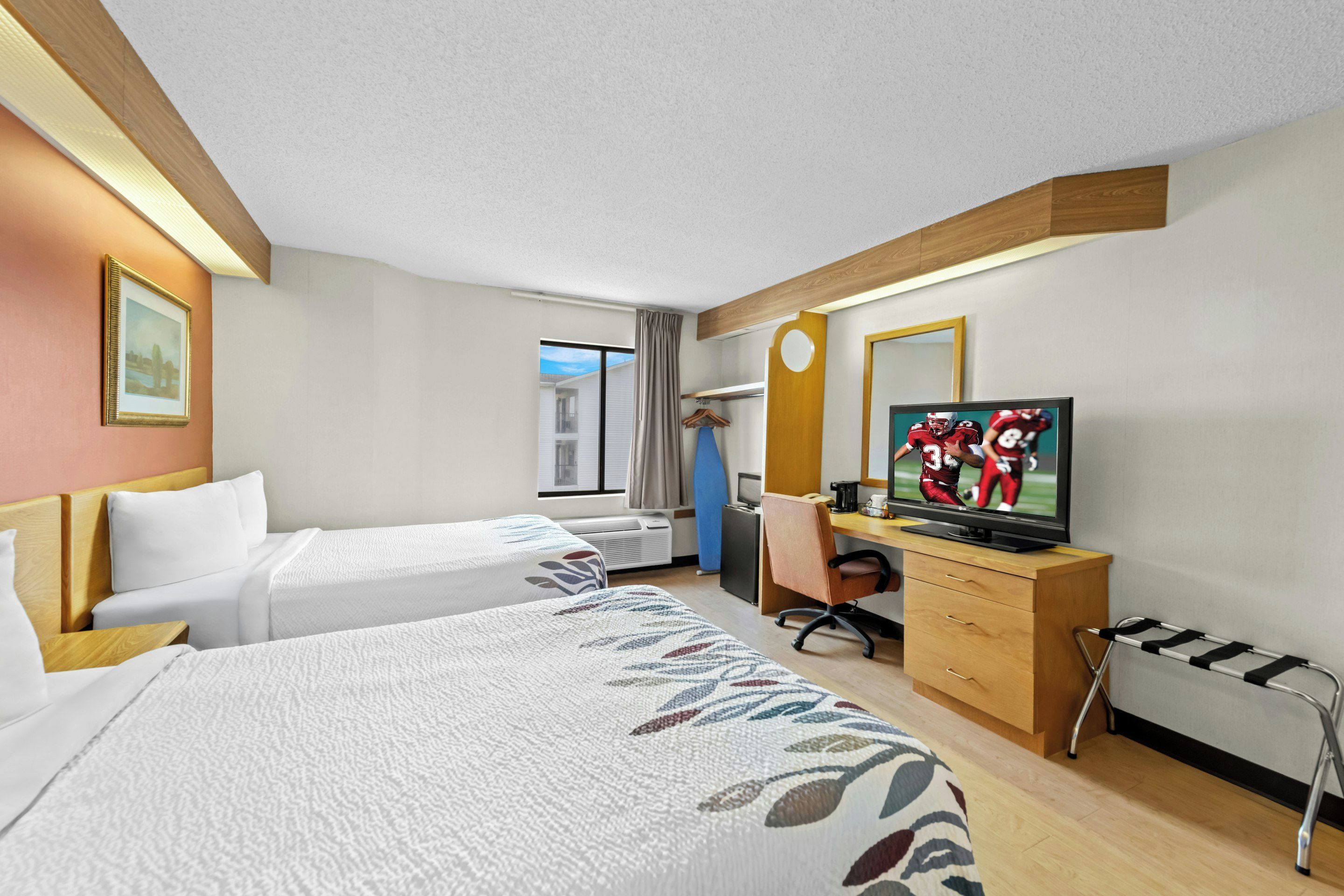 Habitación de hotel con dos camas, un escritorio con televisor, una silla, ventana con cortinas y techo de madera.