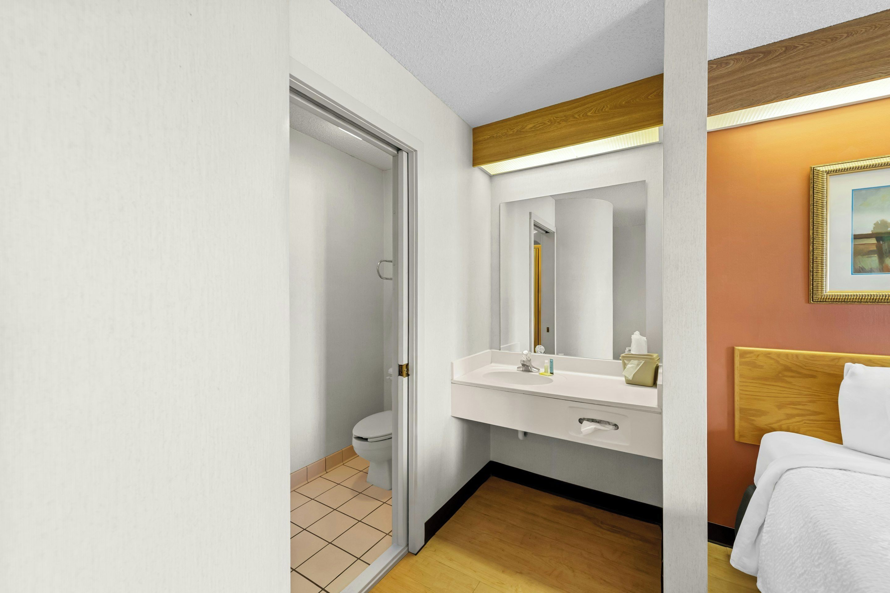 A hotel bathroom with a toilet, sink, mirror, and a bed with a wooden headboard.