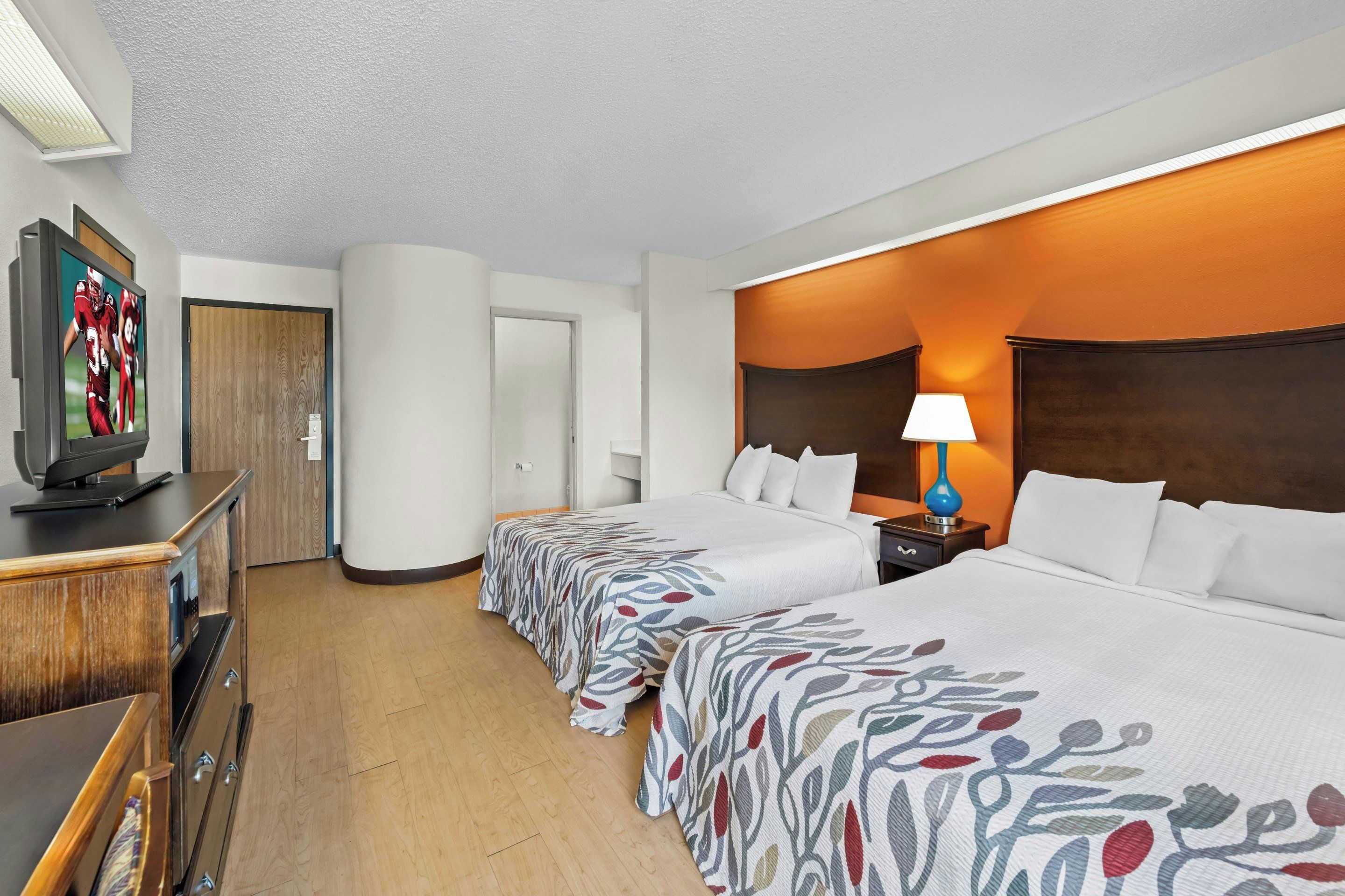 A hotel room with two beds, a TV, a wooden dresser, and a nightstand with a lamp.