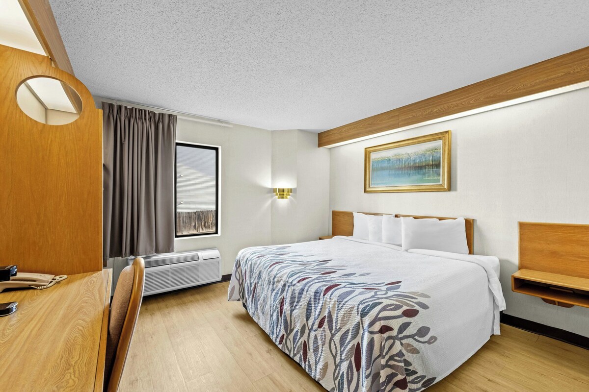 A hotel room with a bed, bedside table, desk, chair, window with curtains, wall-mounted lamp, and a painting on the wall.