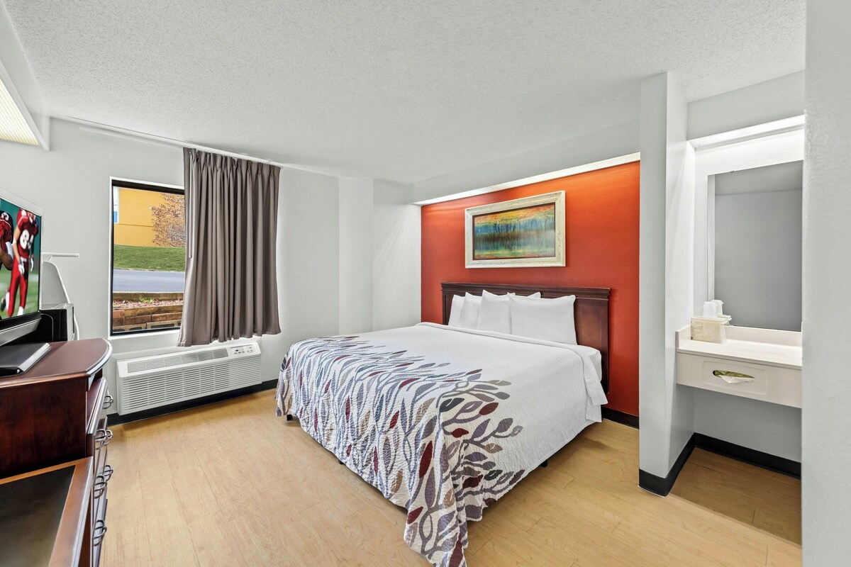 A hotel room with a bed, a desk, a TV, a window with curtains, a painting on the wall, and a bathroom mirror.