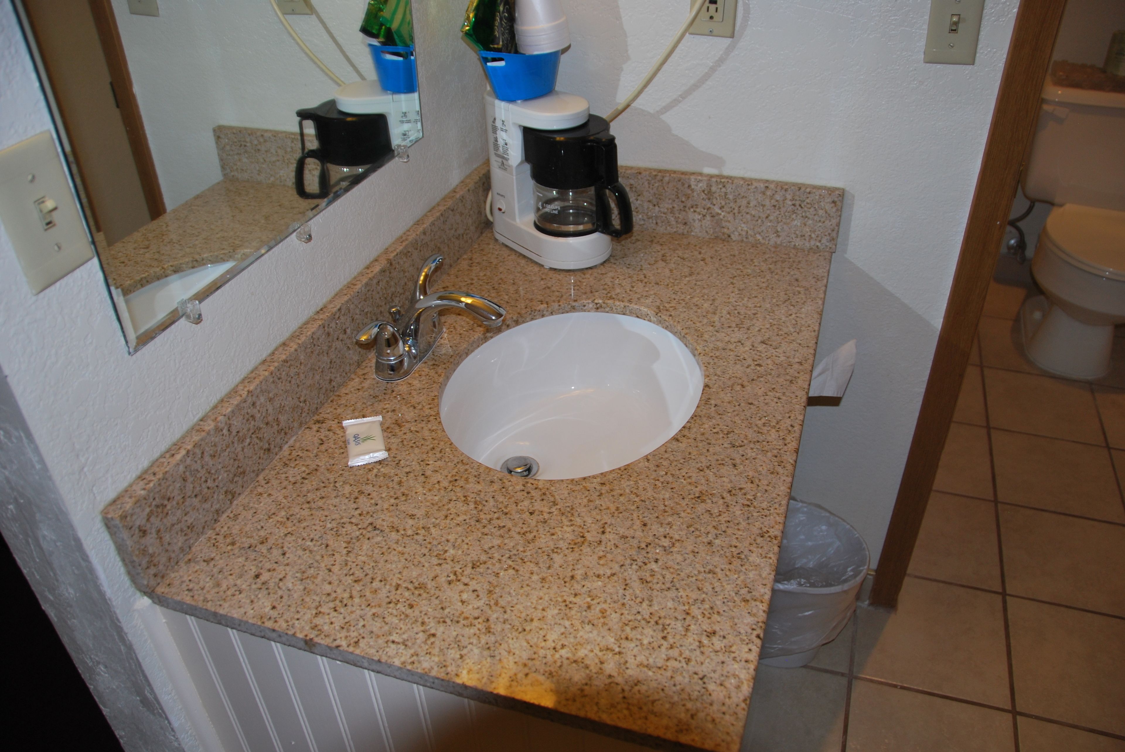 Un baño con encimera de granito, un lavamanos blanco, un espejo, una cafetera y un cubo de basura.