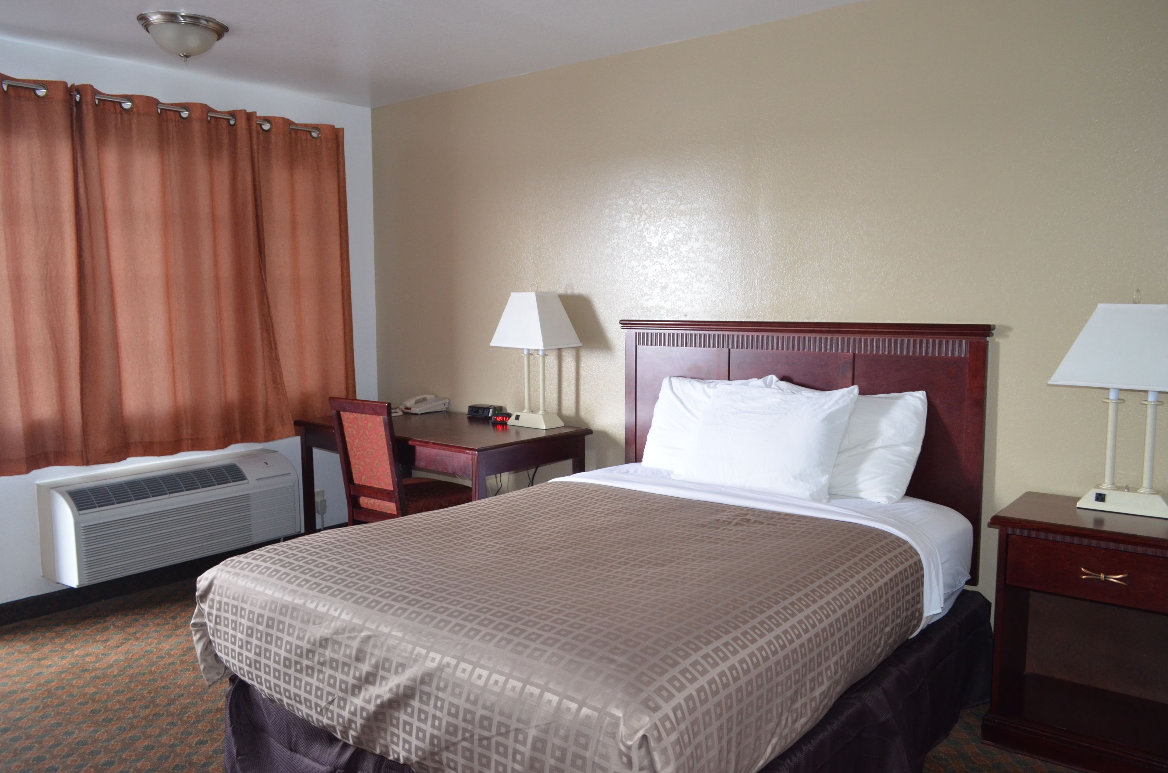 A hotel room with a bed, a desk, a chair, a lamp, and an air conditioning unit.