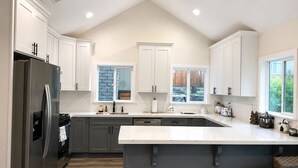 A kitchen with white cabinets, a stainless steel refrigerator, a large island, and a view of the outdoors through windows. - Bear Creek Resort (Big Bear Lake)