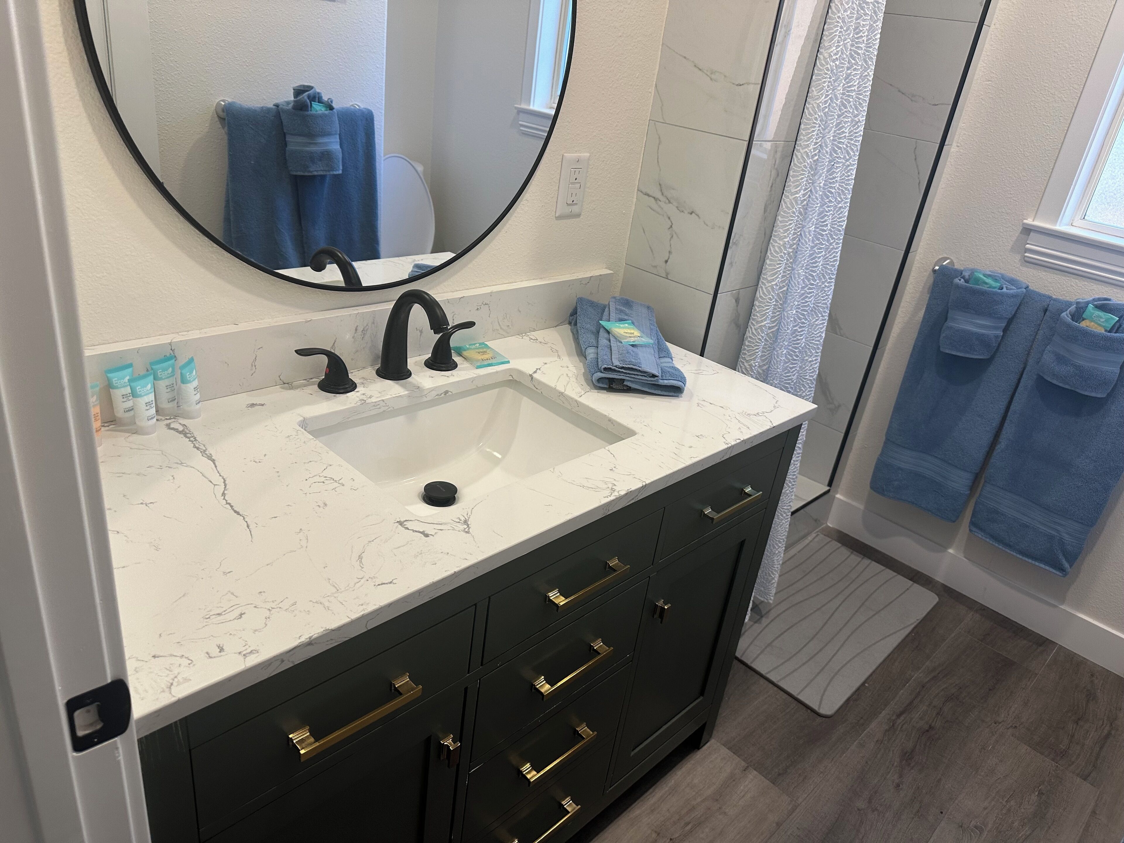 Un baño con lavamanos de mármol blanco, grifos negros y un mueble oscuro con tiradores dorados.