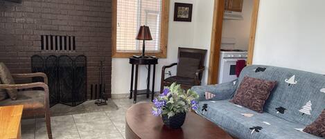 A living room with a brick wall, a sofa, a wooden coffee table, a TV, and a window with blinds.