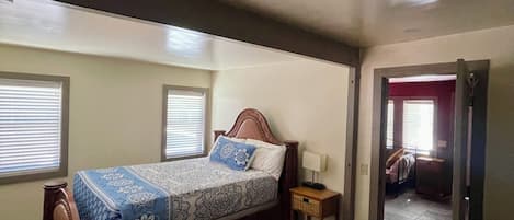 A bedroom with a bed, two nightstands, a door leading to another room, and a window with blinds.