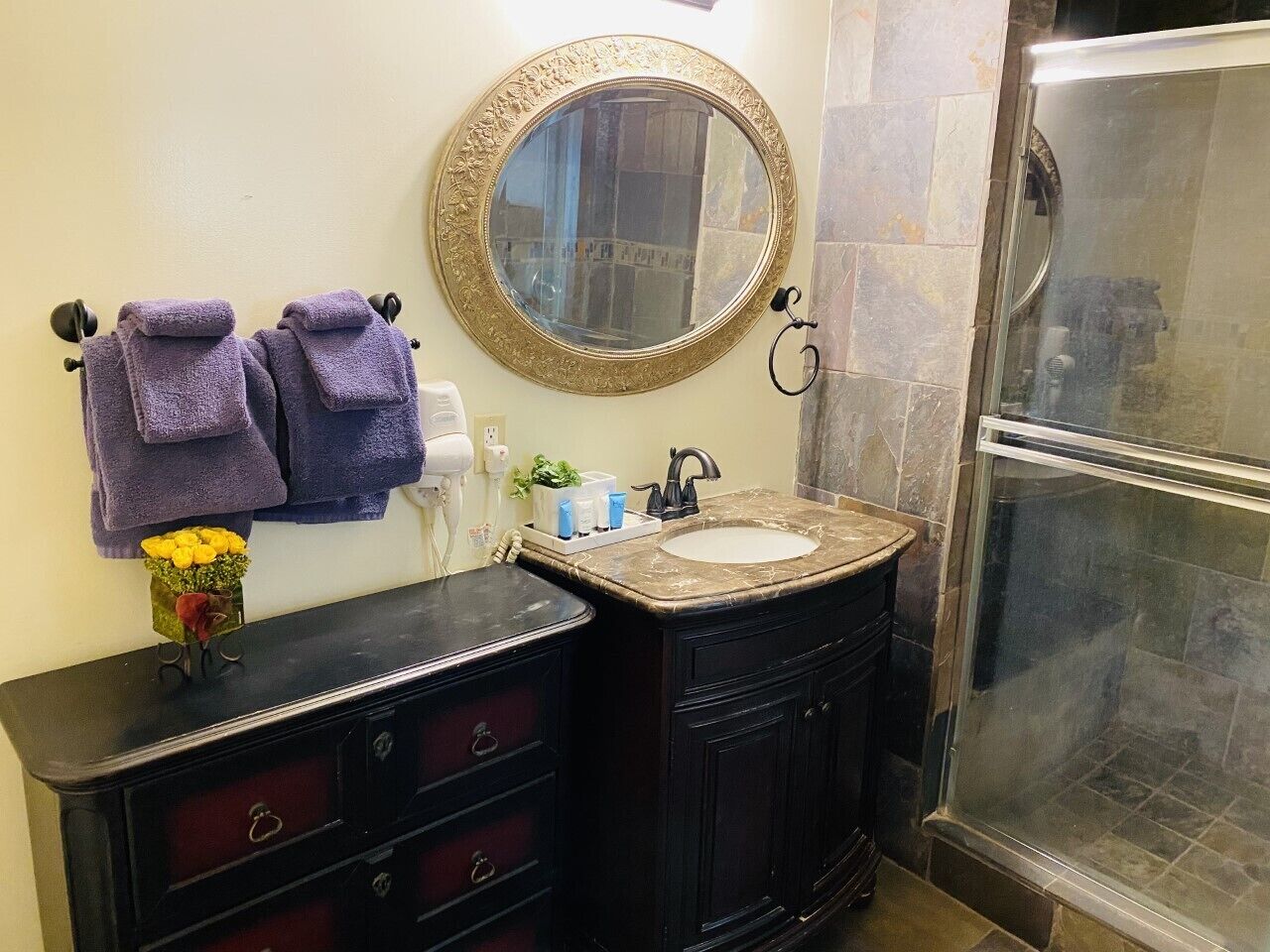 Un baño con lavamanos de mármol, muebles oscuros, un espejo redondo con un marco decorativo y una cabina de ducha de vidrio.