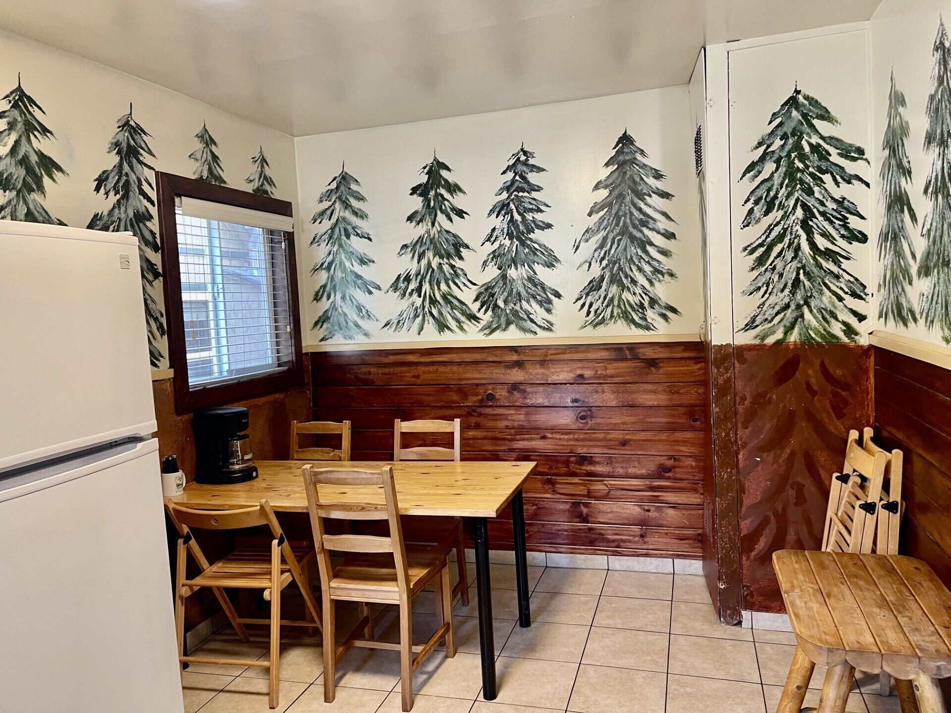 A dining area with a wooden table and chairs, a refrigerator, and a wall with painted pine trees.