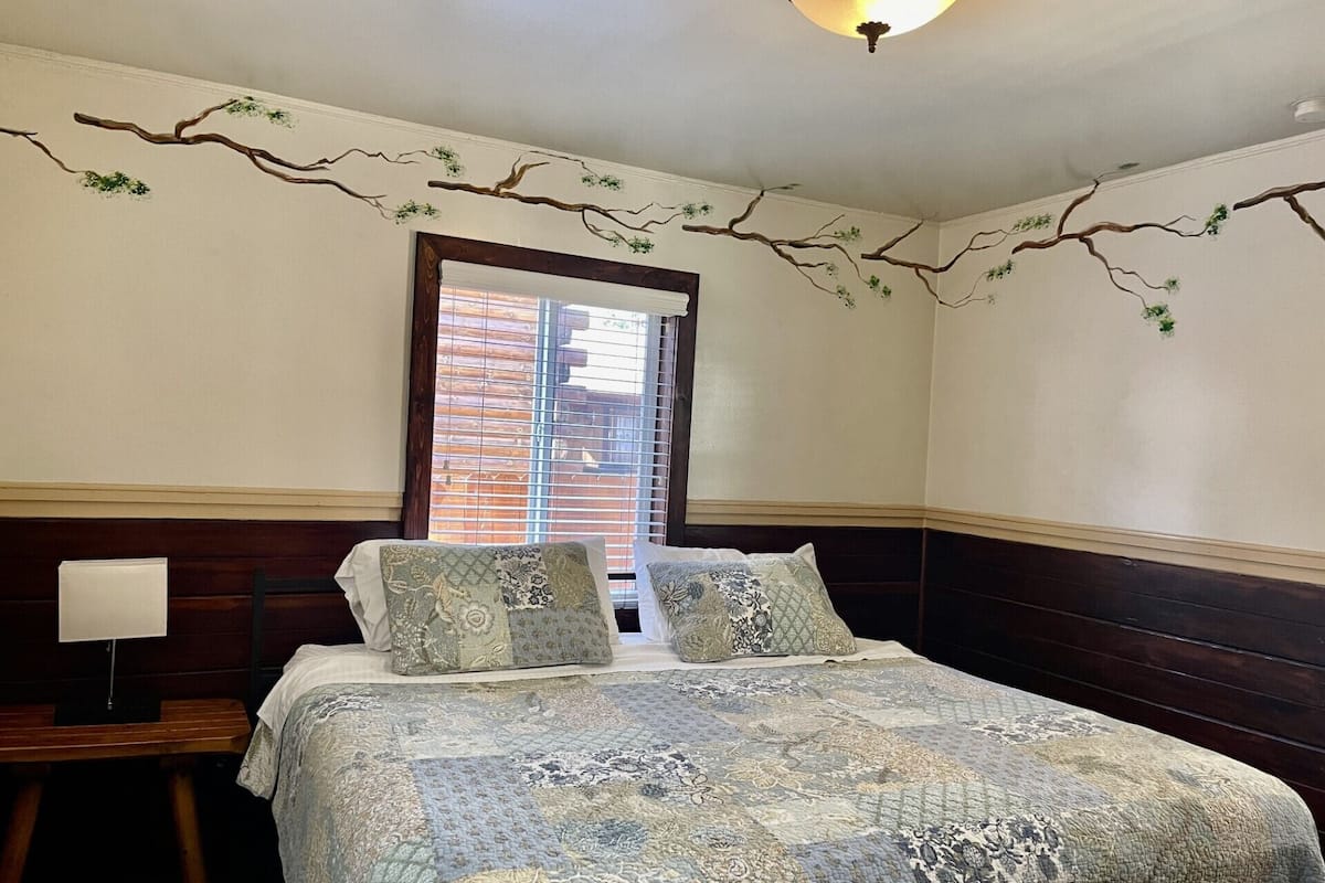 A bedroom with a large bed, two pillows, a nightstand, and a window with blinds.