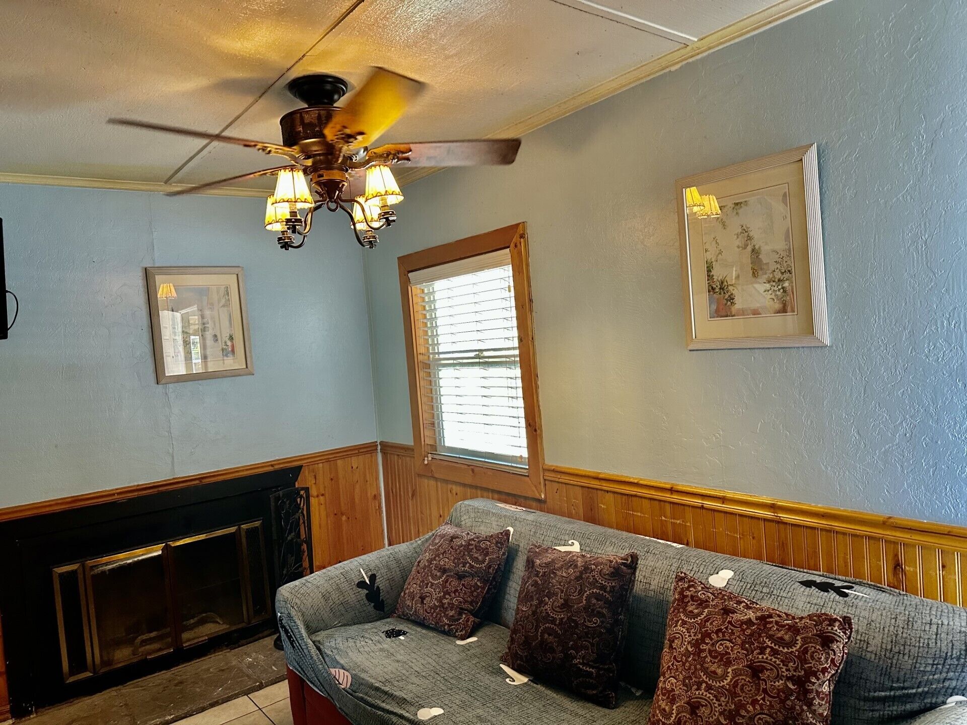 A living room with a fireplace, a sofa with patterned cushions, a ceiling fan, and wall-mounted artwork.