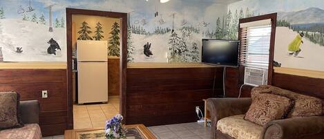 Una sala de estar con techo de madera, un ventilador de techo, un televisor, un sofá, una mesa de centro con un florero y un refrigerador.
