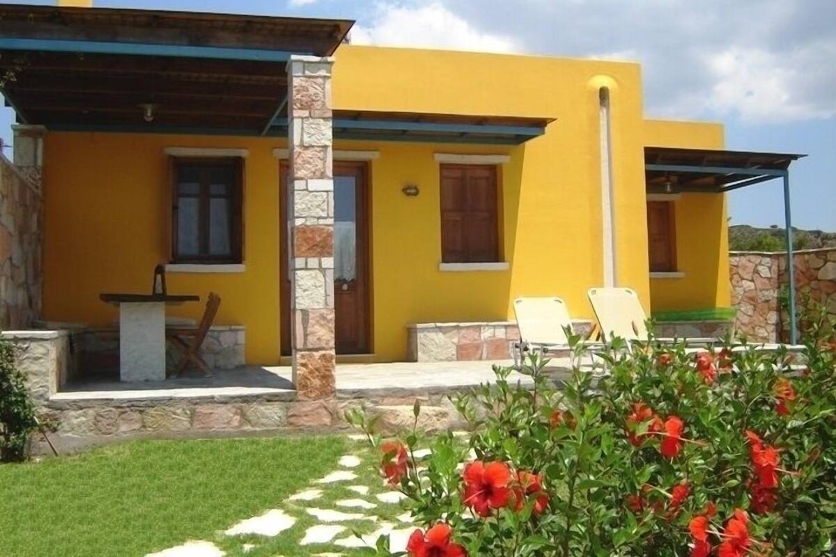 A yellow building with a stone pillar, a door, and a window. There are two white chairs outside.
