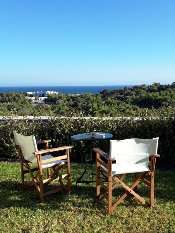 Two wooden chairs and a small table with books on a grassy area overlooking a coastal landscape with buildings in the distance.