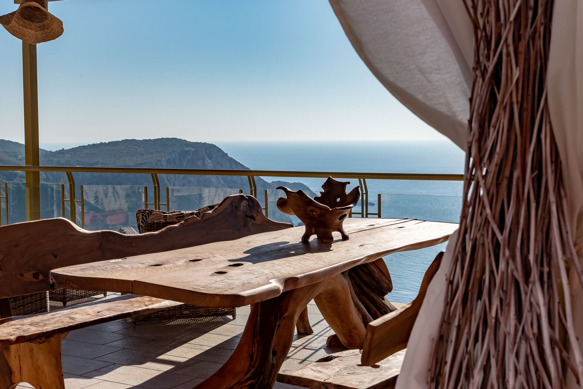 Un tavolo in legno dal design originale, posto su un balcone con vista su un paesaggio montuoso e il mare.