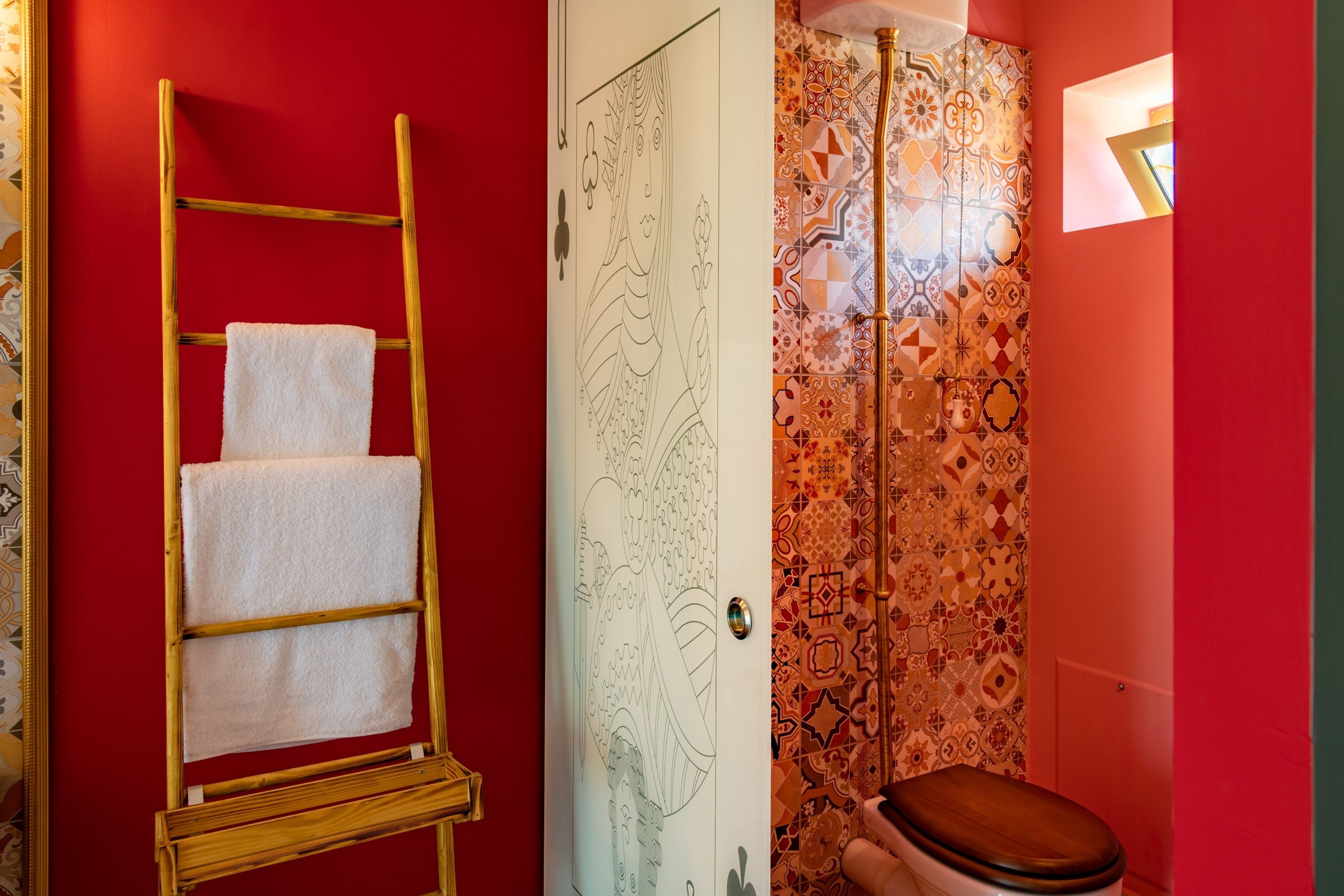 Un baño con pared roja, una escalera de madera con una toalla blanca, una cortina de ducha estampada y un inodoro de madera.
