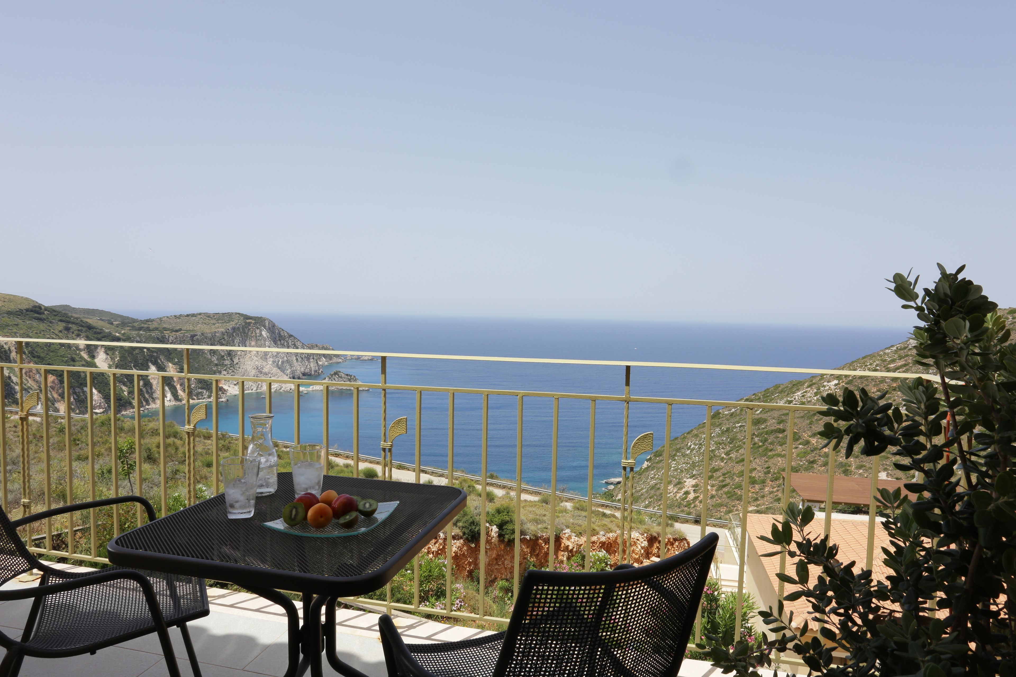 A balcony with a table and chairs, overlooking a coastal landscape with the sea and hills.