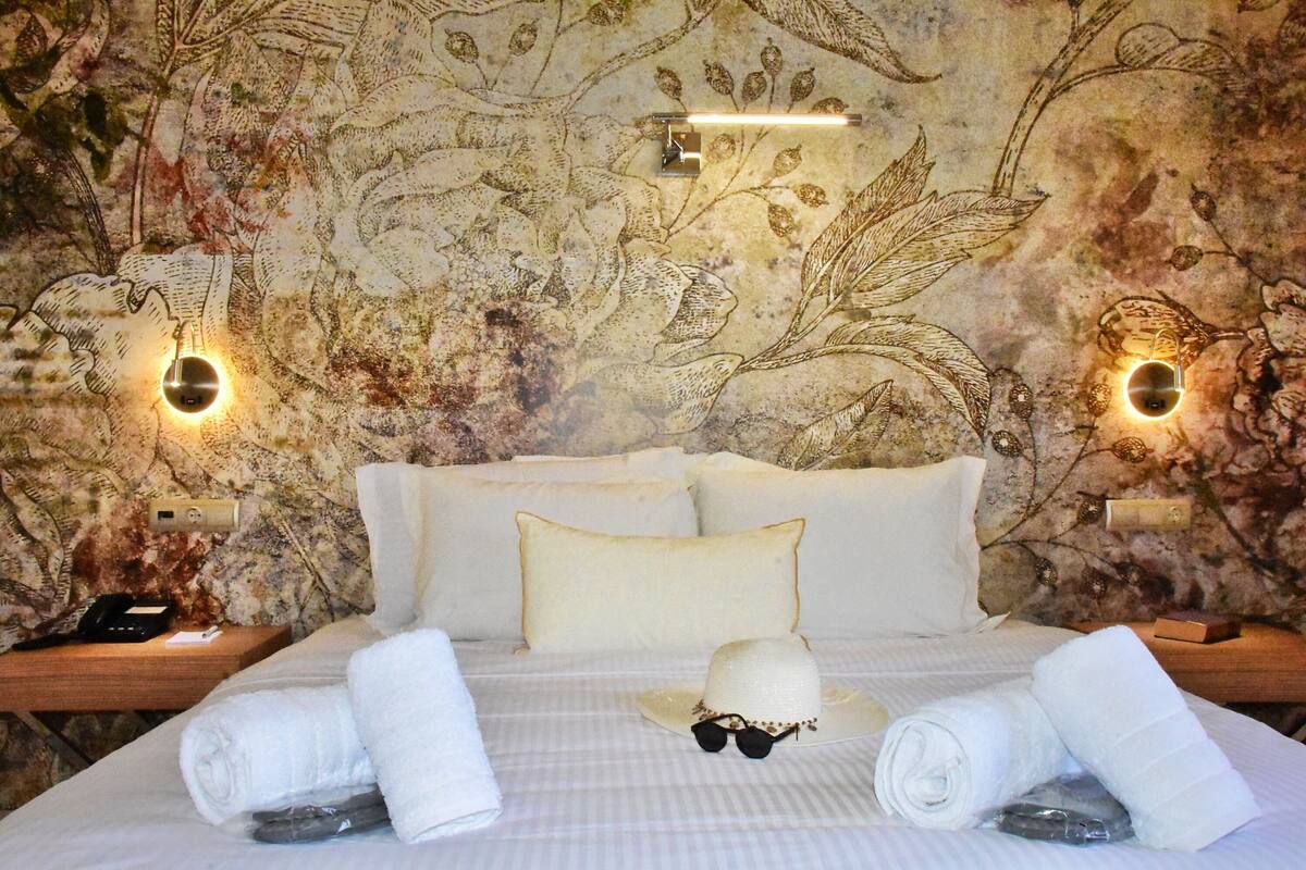 Een hotelkamer met bloemenbehang, een bed met witte lakens, twee wandlampen en een nachtkastje met een telefoon en een doos.