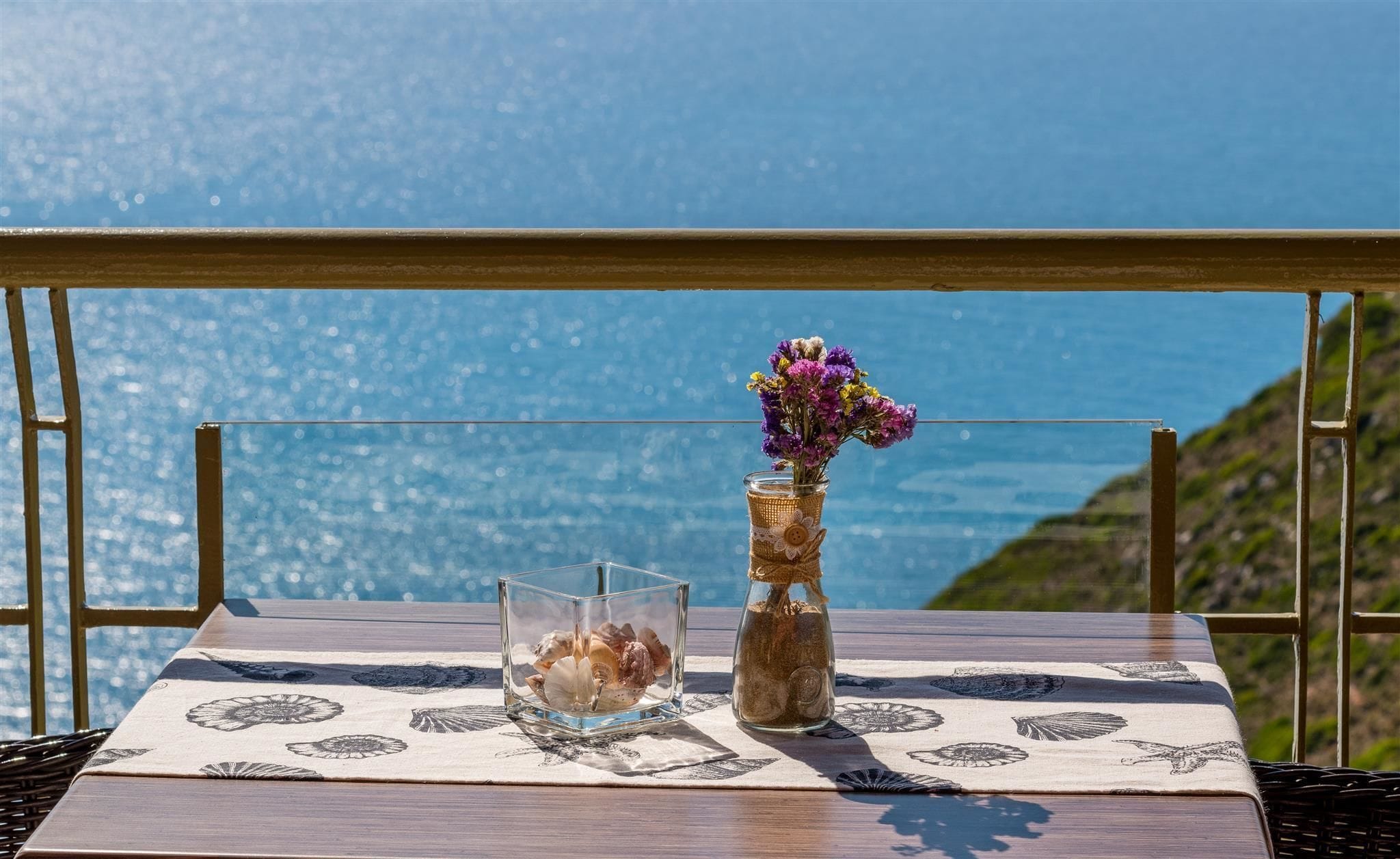A table with a vase of flowers and a glass container of seashells on a balcony overlooking a body of water.