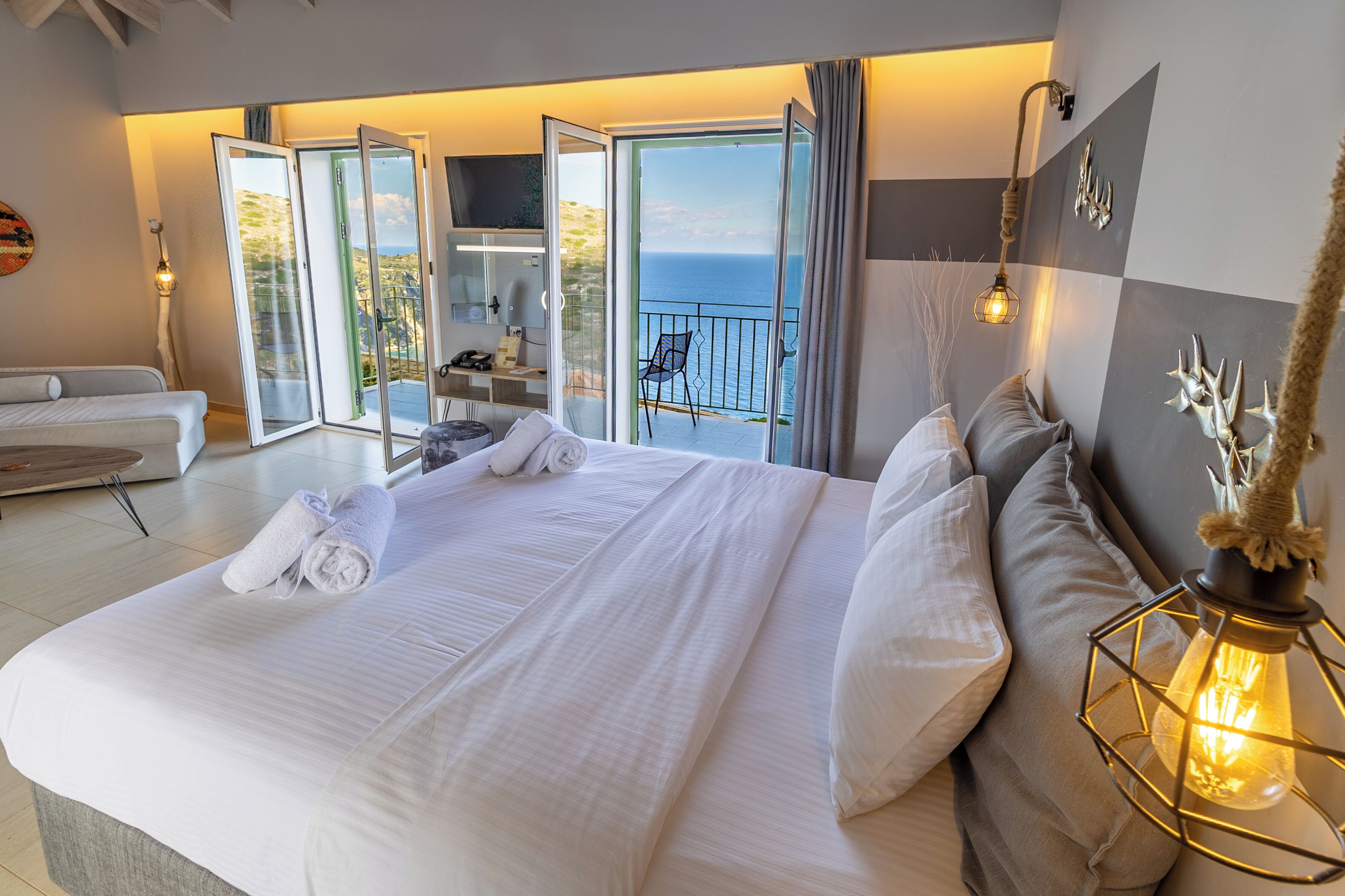 A hotel room with a large bed, a balcony with a view of the sea, a desk with a chair, and a TV.