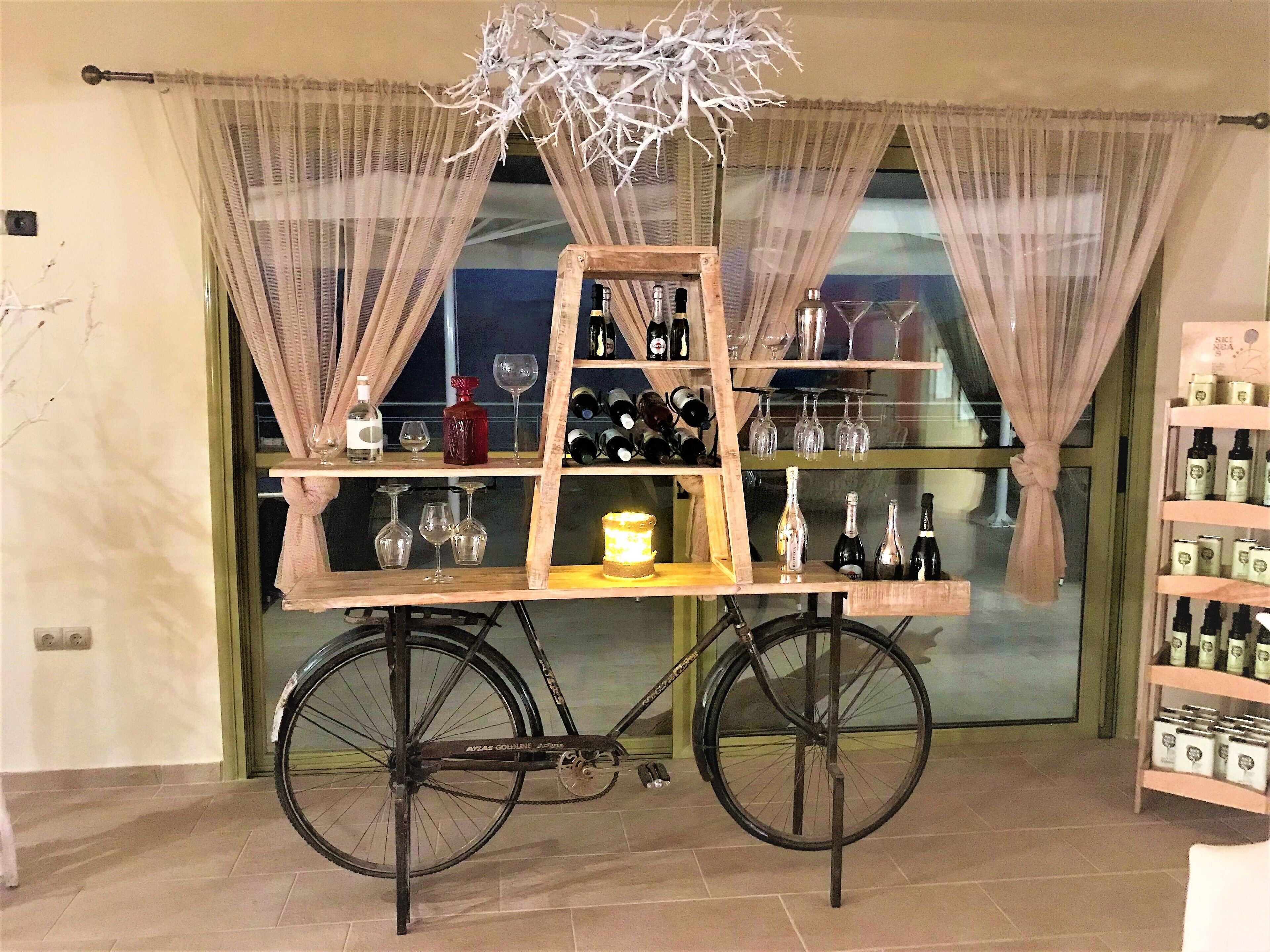 A bicycle cart with shelves holding bottles and glasses, positioned in front of a window with sheer curtains.