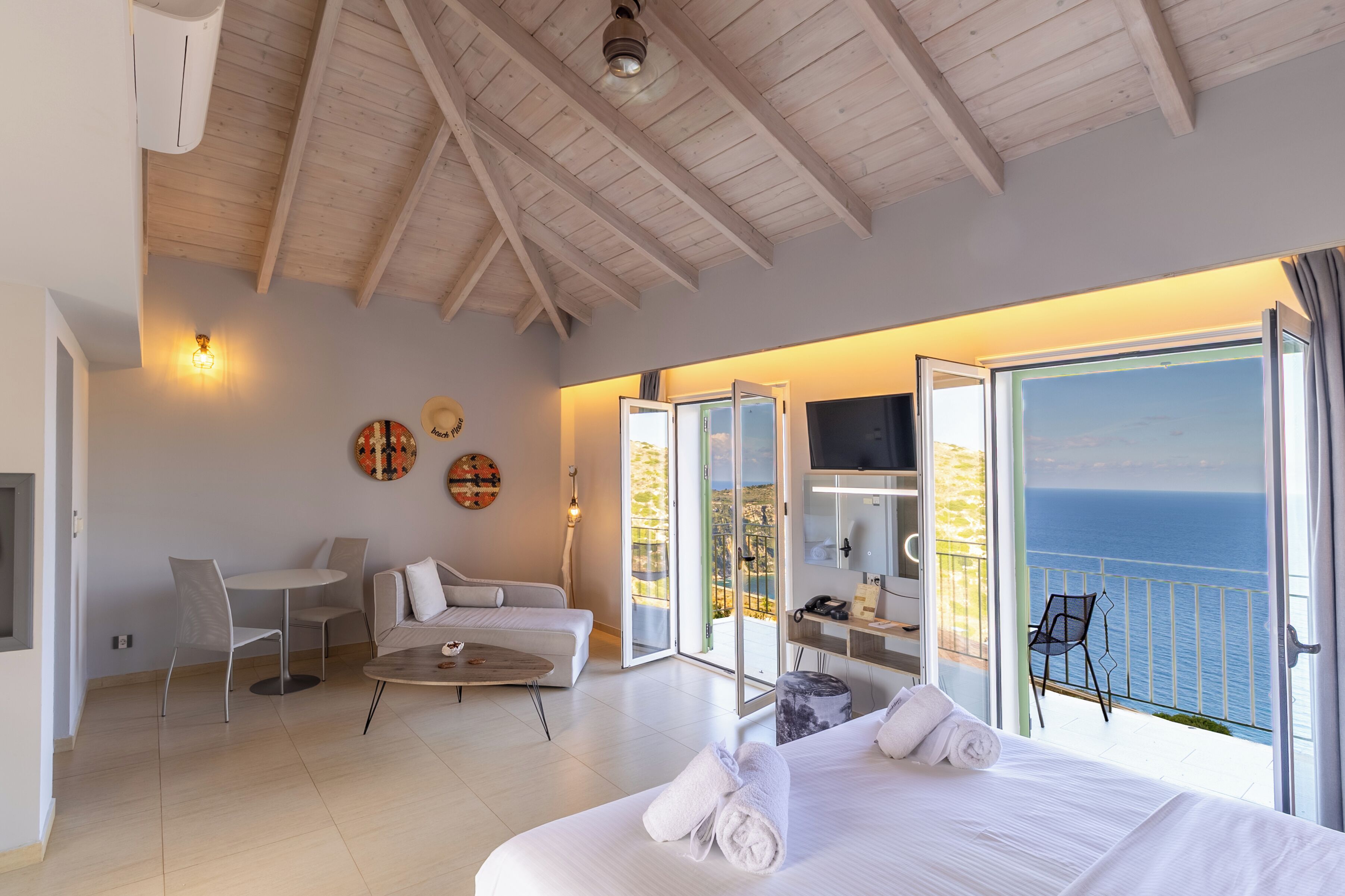 A spacious bedroom with a large bed, a desk, and a chair. There is a balcony with a view of the sea.
