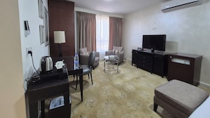 A hotel room with a sofa, armchair, TV, and a desk with a kettle and water bottle. - Le Vendôme Hotel (Amman)