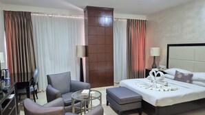 A hotel room with a bed, two armchairs, a small round table, a desk with a lamp, and a large window with curtains. - Le Vendôme Hotel (Amman)