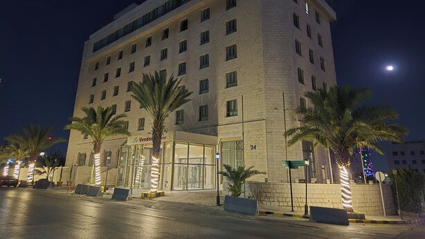 A multi-story hotel with a modern design, featuring a large entrance with glass doors and palm trees. - Le Vendôme Hotel (Amman)