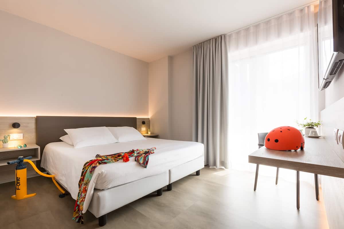 Moderni hotellihuone, jossa on sänky, yöpöytä, lamppu ja kypärä pöydällä.