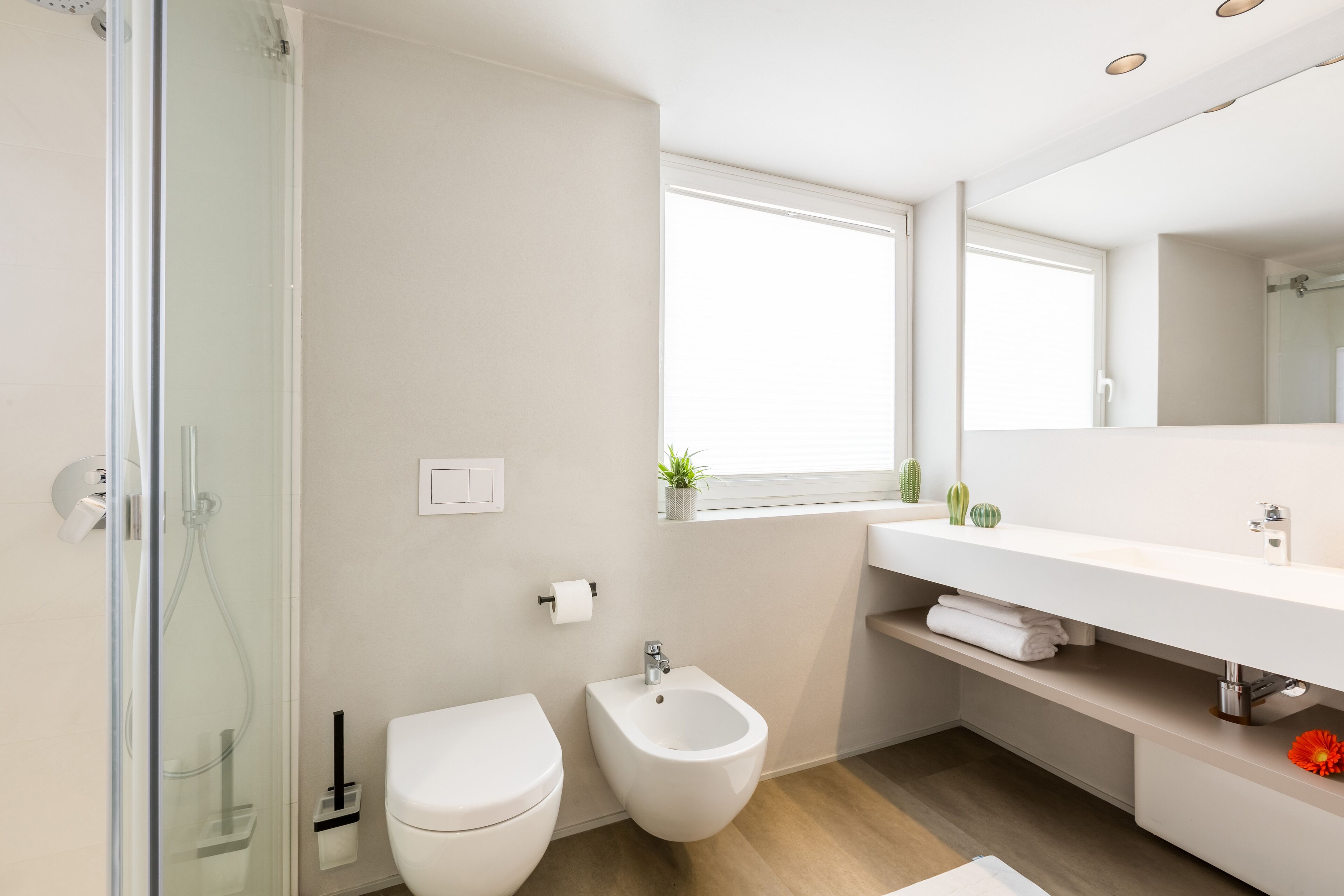 A modern bathroom with a glass shower enclosure, a toilet, a bidet, a vanity with a mirror, and a window providing natural light.