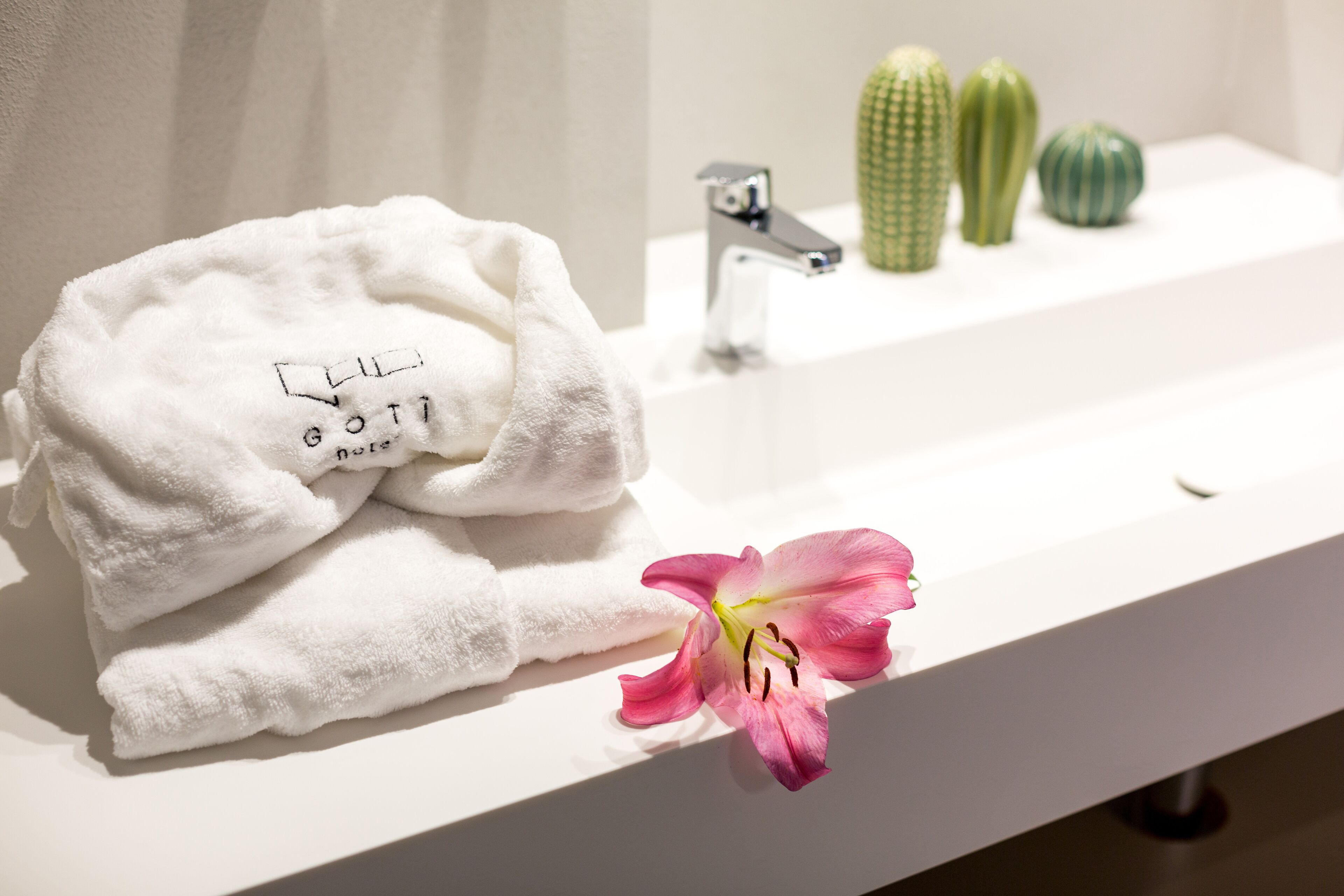 A white towel with text, a pink flower, and three green cacti on a white bathroom counter.