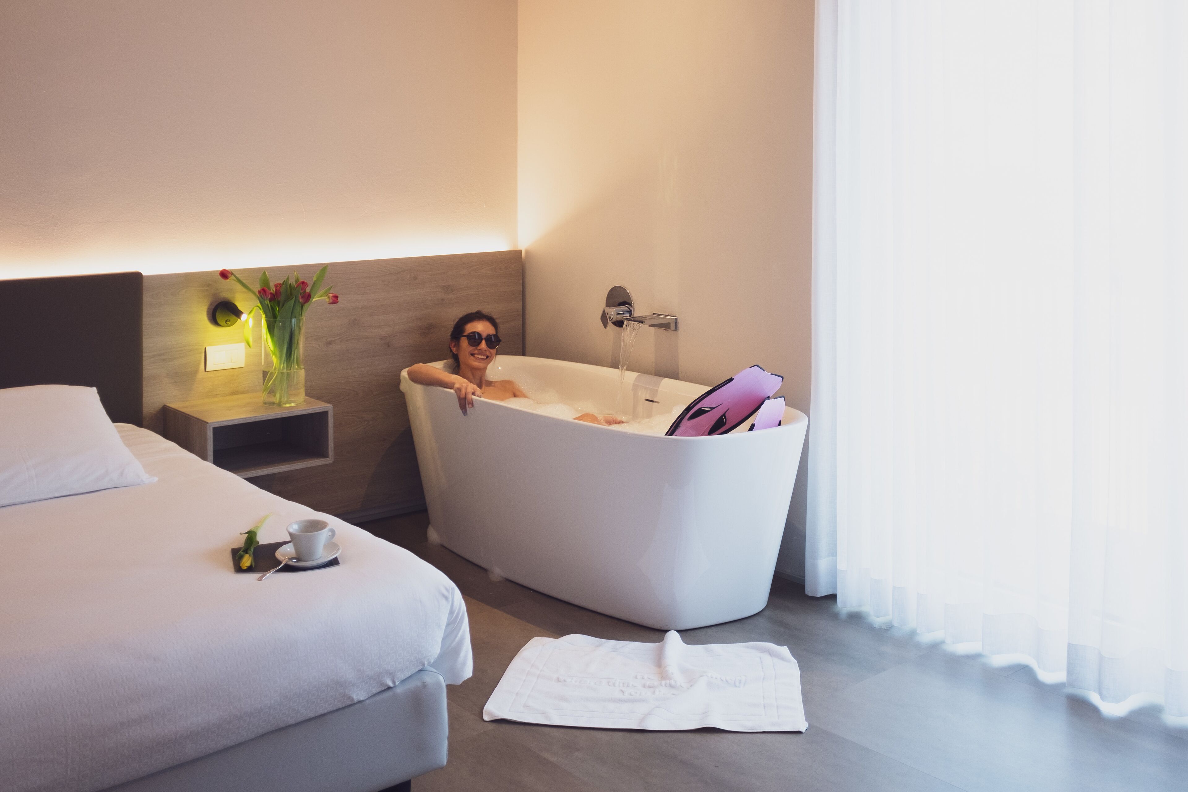 Ein modernes Hotelzimmer mit einer großen Badewanne, einem ordentlich bezogenen Bett, einem Nachttisch mit Blumen und einer Tasse Kaffee auf dem Bett.
