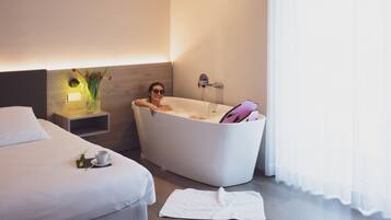Ein modernes Hotelzimmer mit einer großen Badewanne, einem ordentlich bezogenen Bett, einem Nachttisch mit Blumen und einer Tasse Kaffee auf dem Bett.