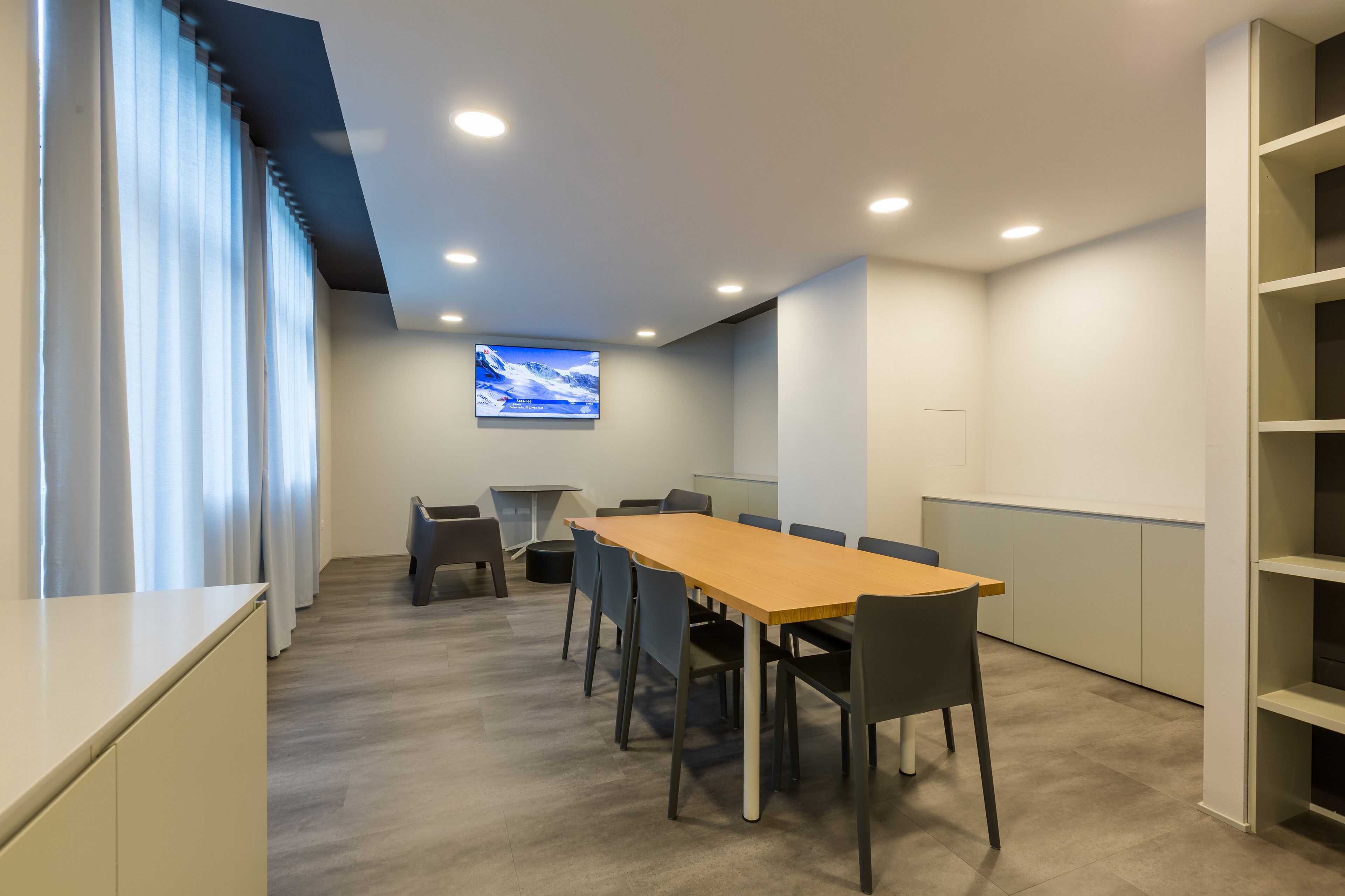 A conference room with a long table, chairs, a TV screen, and a window with curtains.