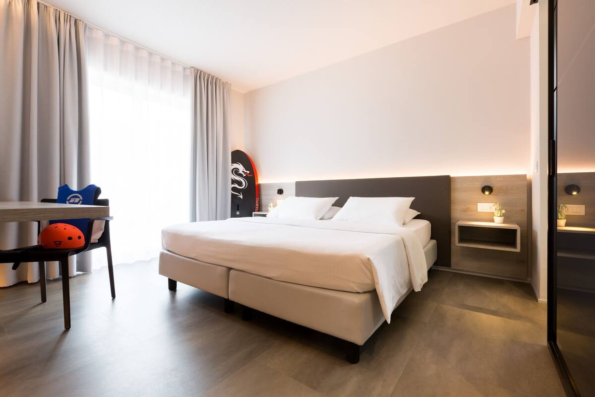 Moderni hotellihuone, jossa on suuri sänky, yöpöydät, työpöytä tuolilla ja ikkuna, jossa on verhot.