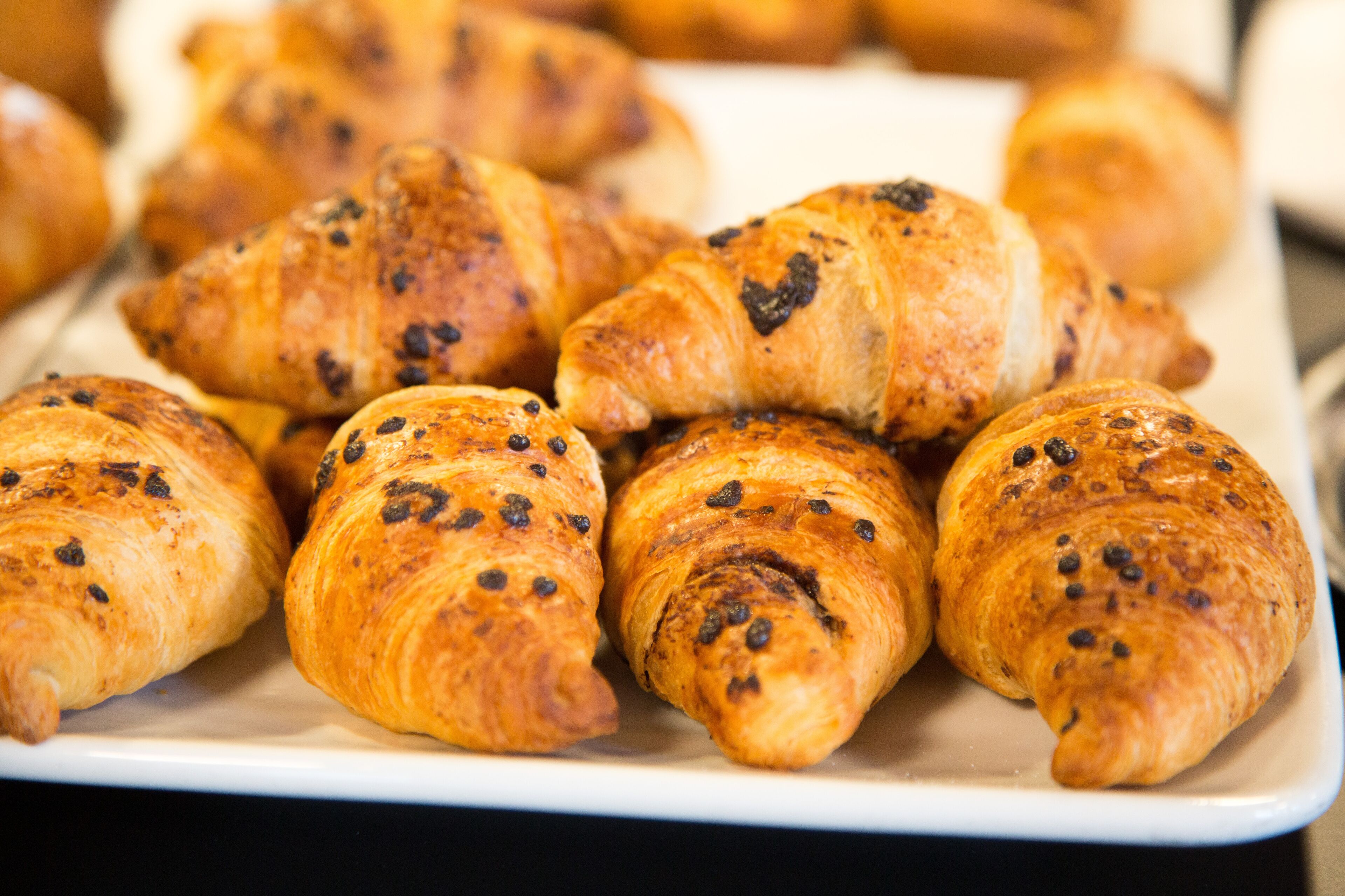 Un plat rectangular blanc amb una safata de croissants daurats i marrons, amb llavors negres visibles, probablement xocolata o raïm.