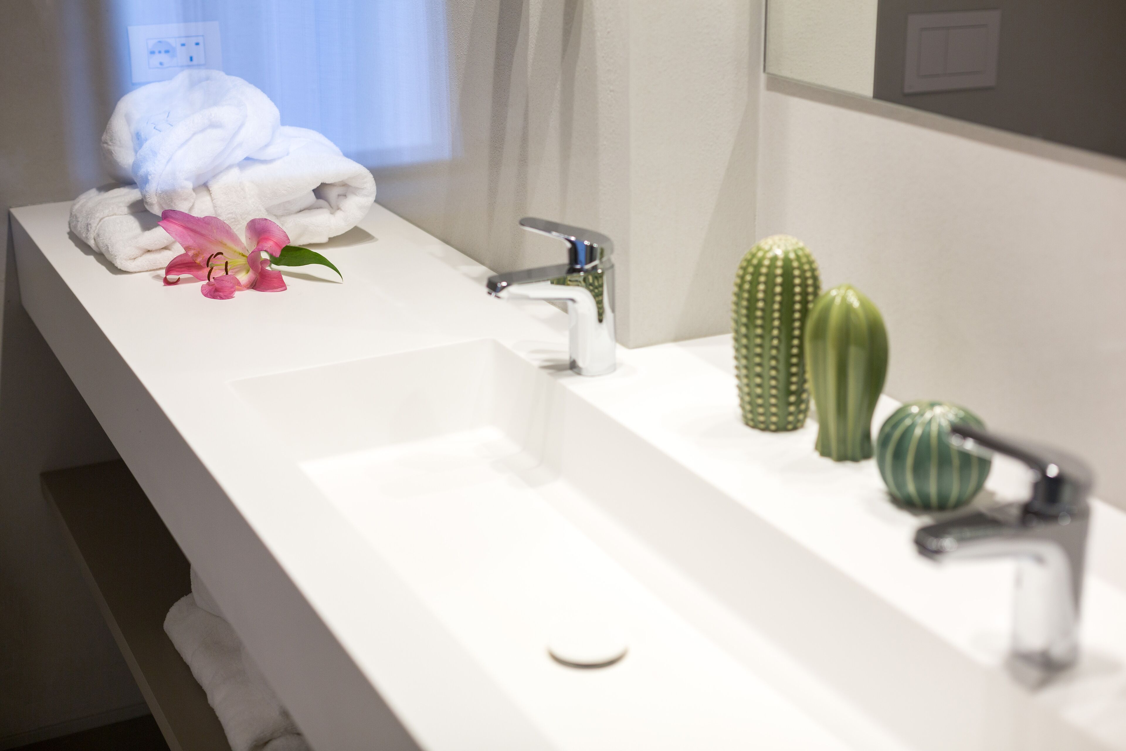Une salle de bain avec un lavabo blanc, deux robinets, une serviette, une fleur rose et deux cactus verts.
